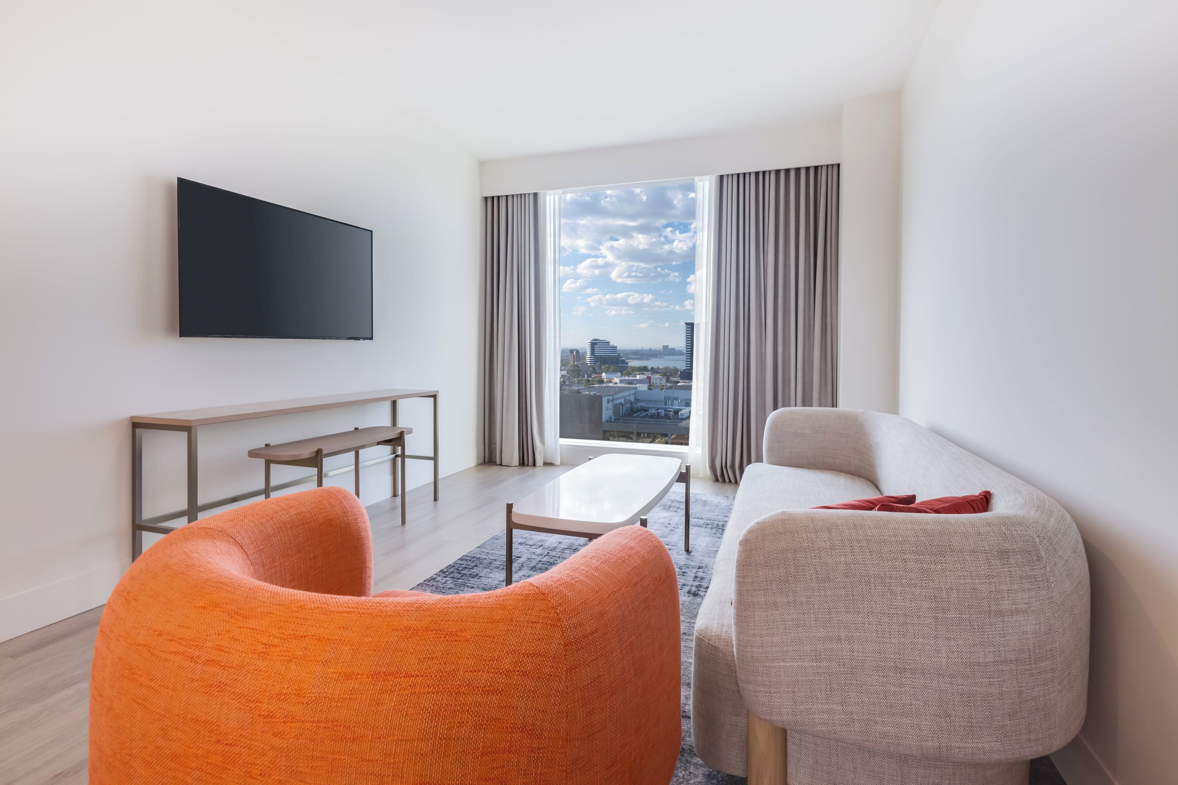 Ein modernes Wohnzimmer mit einem Flachbildfernseher, einer Couch, einem orangefarbenen Sessel, einem kleinen Tisch und Blick auf die Stadt durch große Fenster.