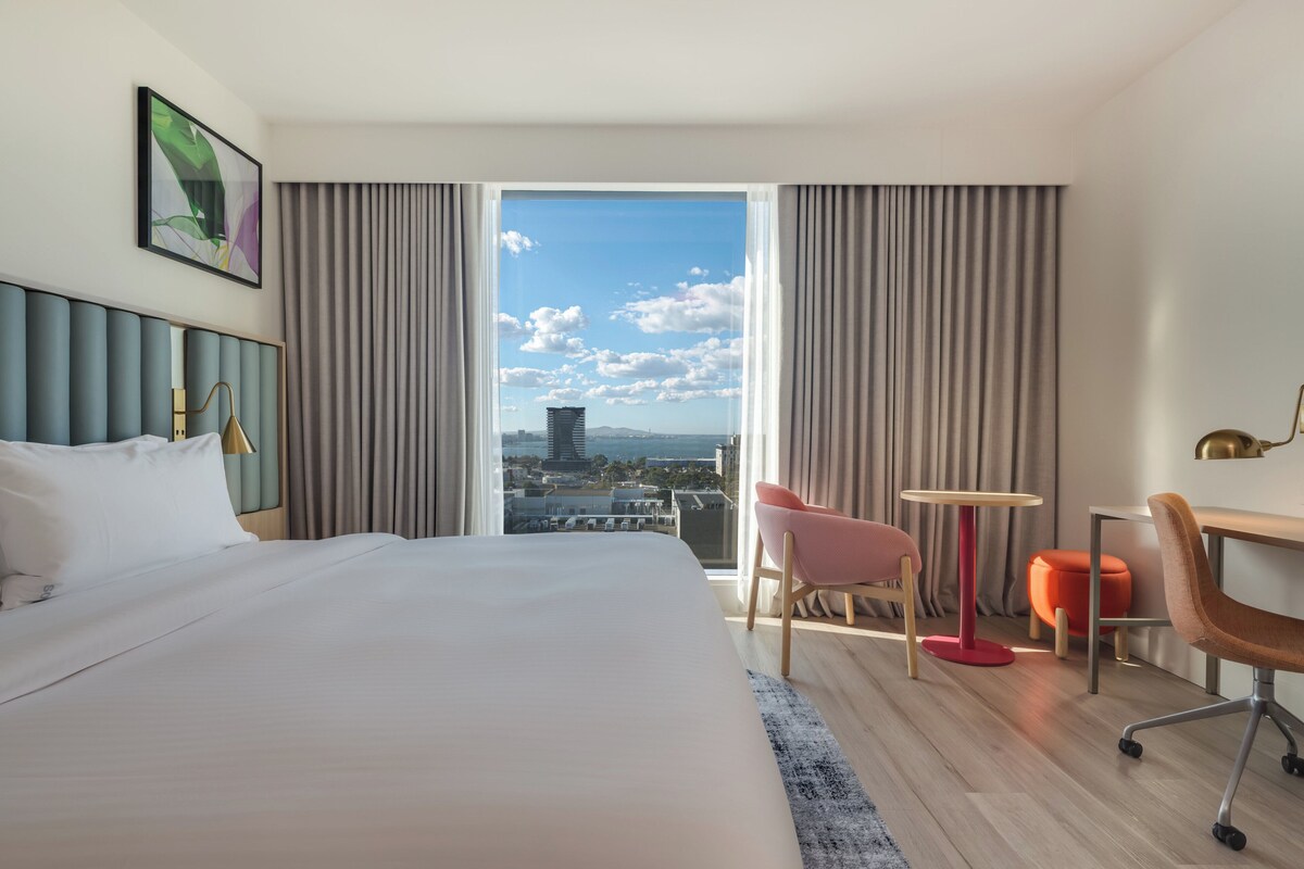 A hotel room with a large bed, a desk, two chairs, a small table, and a view of the city through a large window.