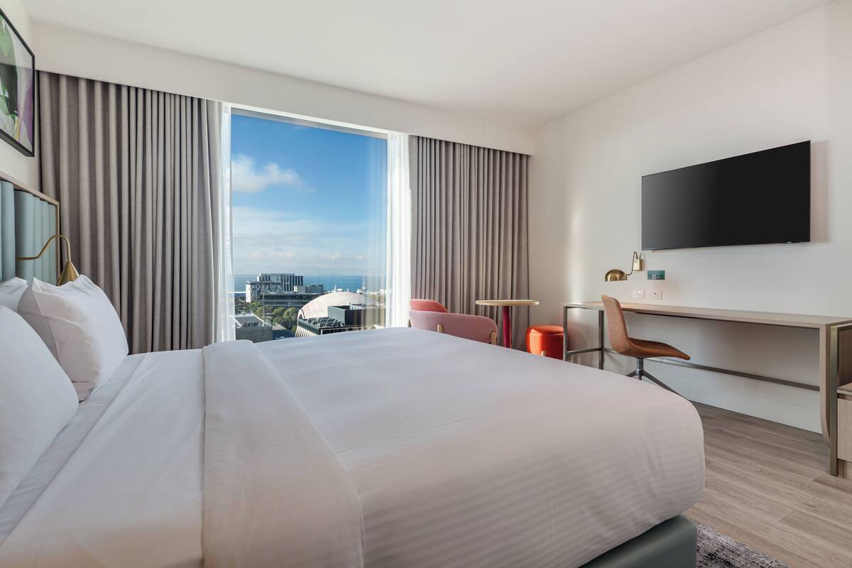 A hotel room with a large bed, a desk, a chair, a television, and a view of the city.