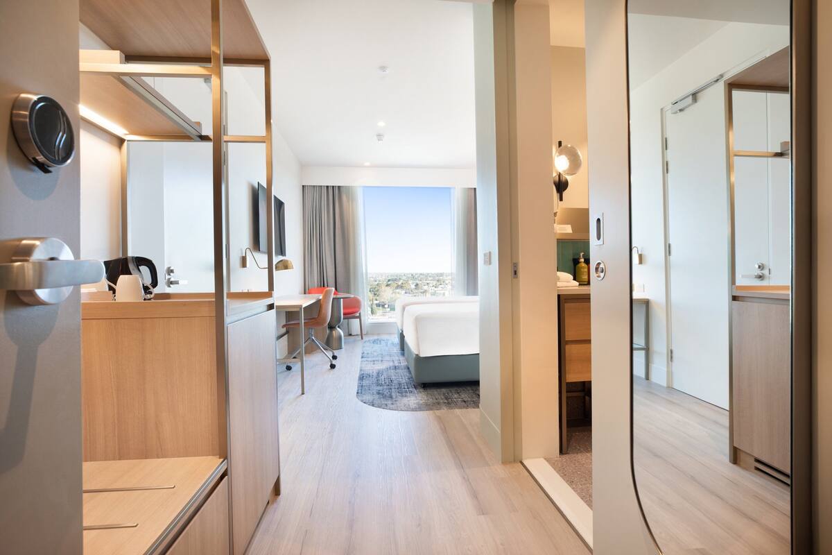 A hotel room with a bed, desk, chair, and a view of the city.