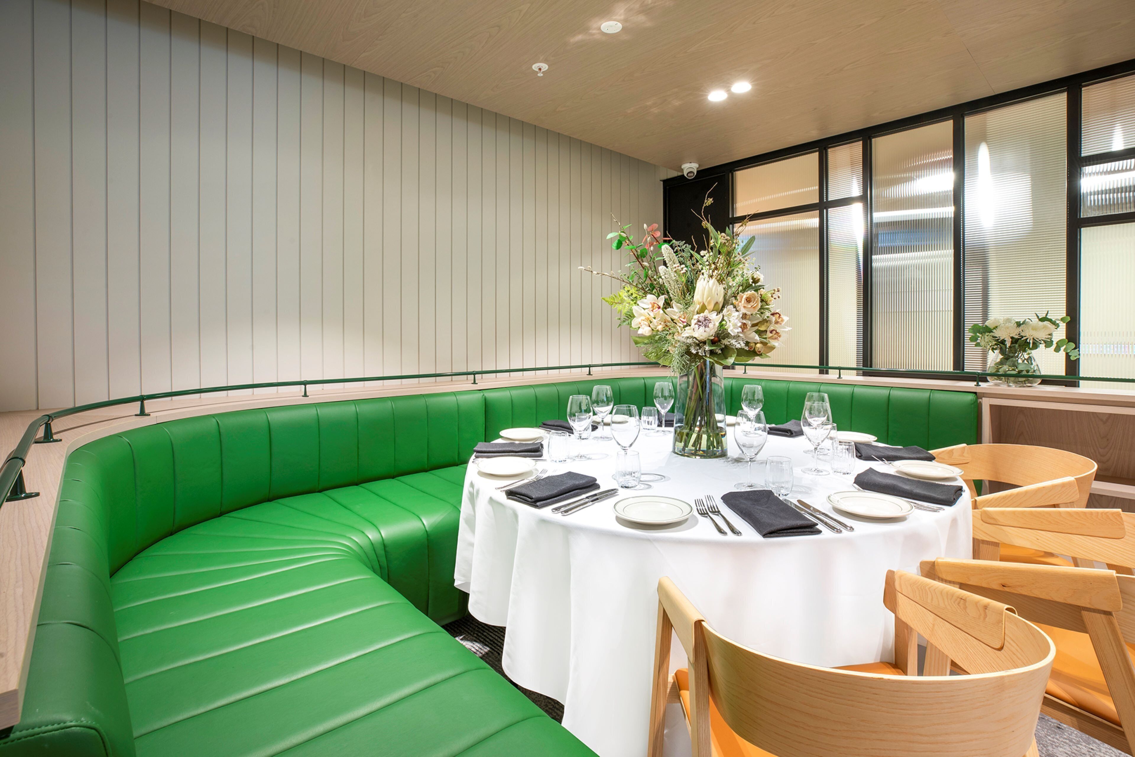 A restaurant with a green booth seating area, round tables with white tablecloths, and a large window with blinds.