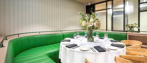 A restaurant with a green booth seating area, round tables with white tablecloths, and a large window with blinds.