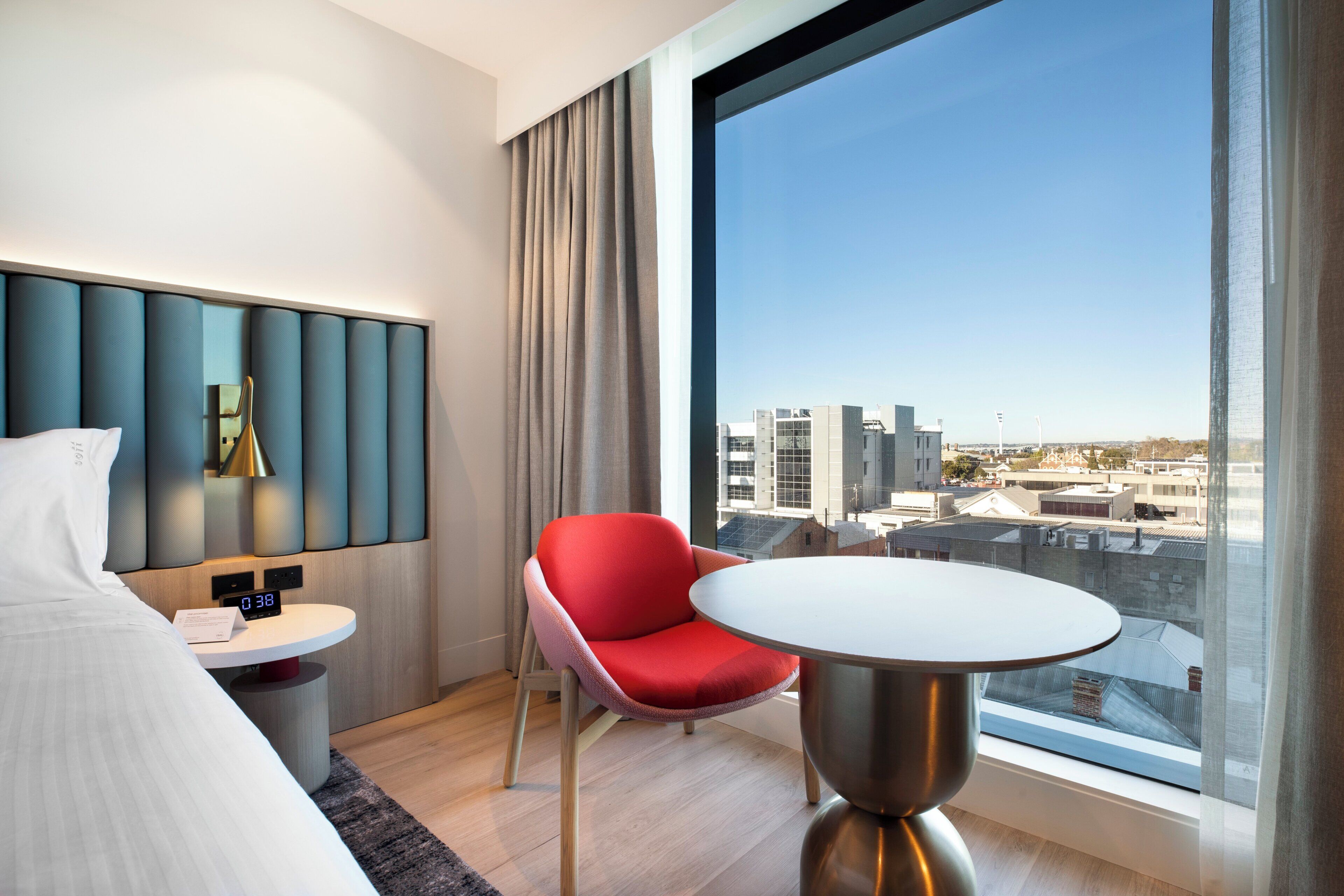Ein modernes Hotelzimmer mit einem Bett, einem roten Sessel, einem runden Tisch, einer digitalen Uhr und einem großen Fenster mit Blick auf die Stadt.