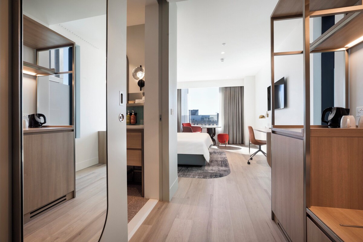 A modern hotel room with a wooden floor, a bed with a white duvet, a desk with a chair, a TV on the wall, and a city view through large windows.