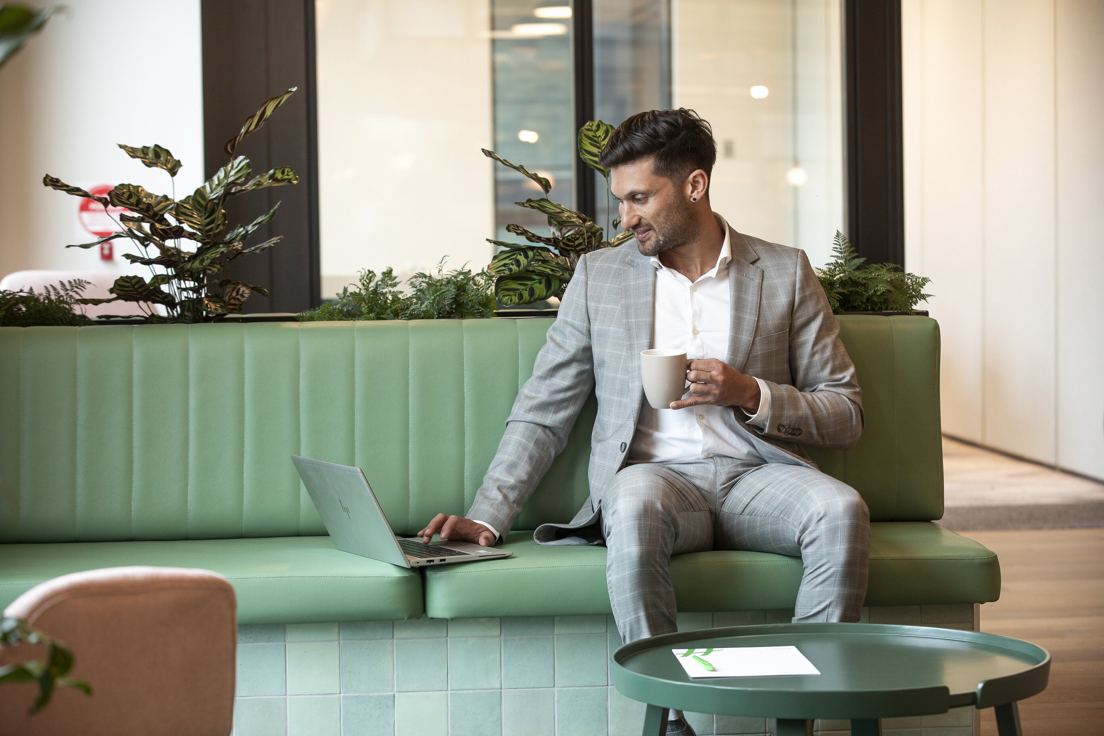 A man in a grey suit sitting on a green couch with a laptop, holding a mug, in a modern indoor setting with plants and a glass wall.