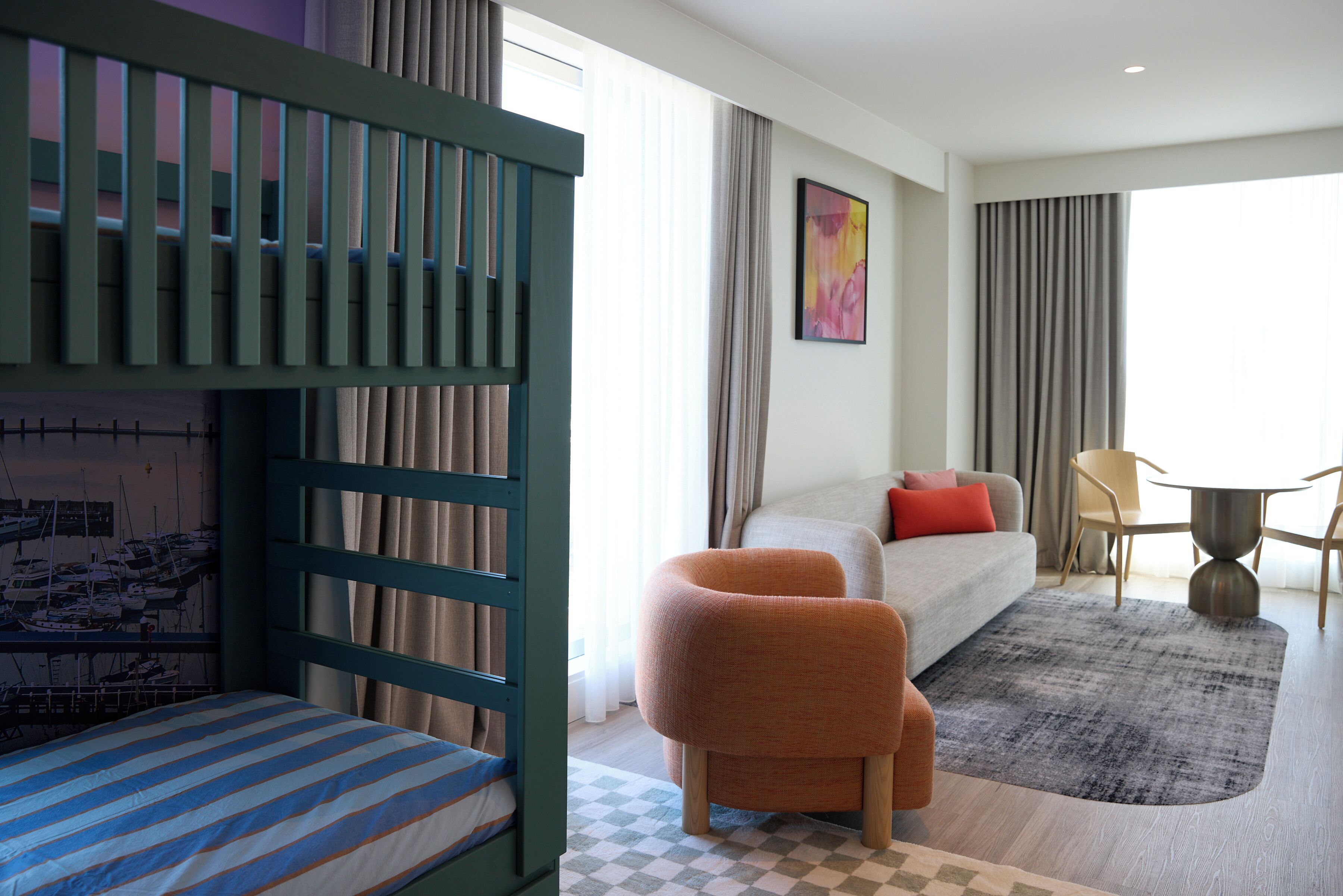 A hotel room with a bunk bed, a sofa, a chair, a dining table, and a painting on the wall.