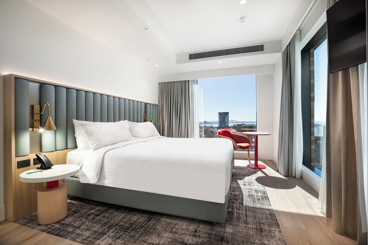 A modern hotel room with a large bed, a small round table, a red chair, and a view of the city through large windows.