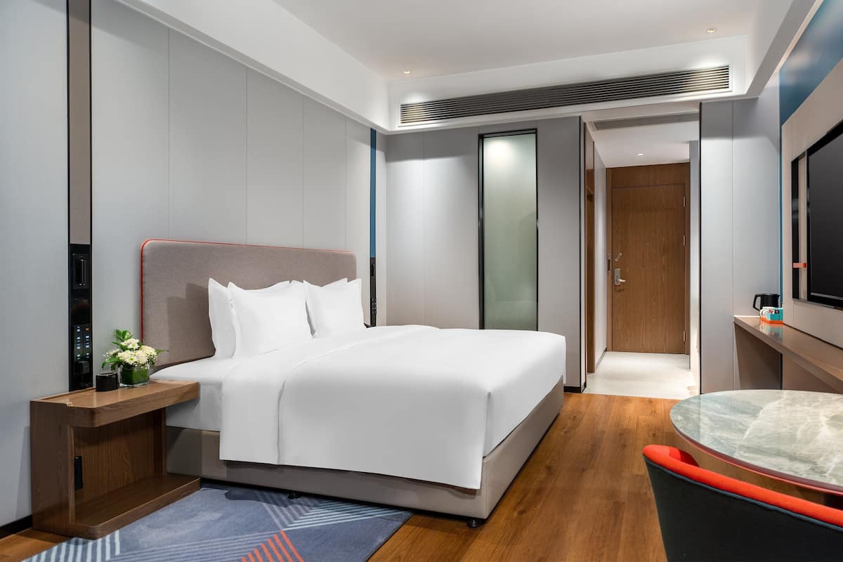 A modern hotel room with a bed, bedside table, TV, and a small table with a chair.