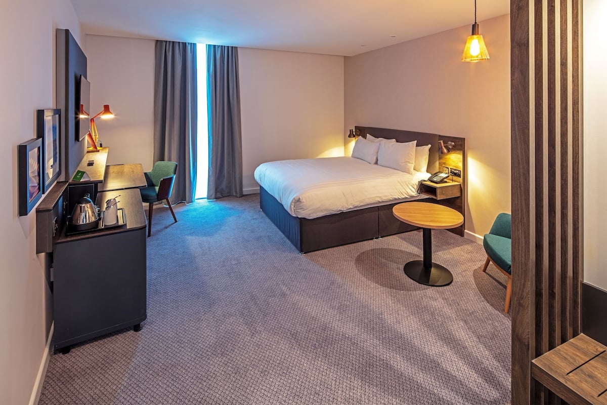 A modern hotel room with a large bed, a desk with a chair, a small round table, and a TV on the wall.