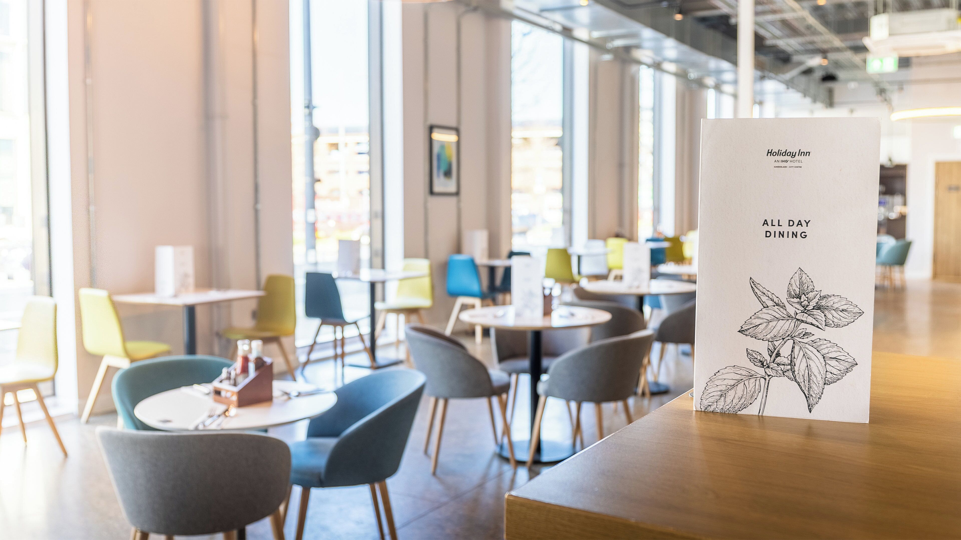 A Holiday Inn All Day Dining menu with a leaf illustration, set in a modern dining area with round tables and chairs.