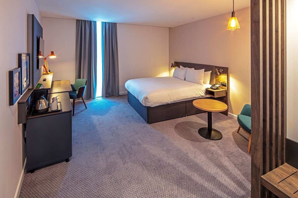 Une chambre d’hôtel moderne avec un grand lit, un bureau avec une chaise, une petite table ronde et un téléviseur fixé au mur.