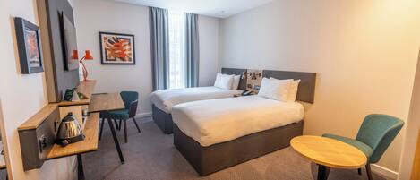 Habitación de hotel con dos camas, un escritorio con silla, una mesita y una lámpara.