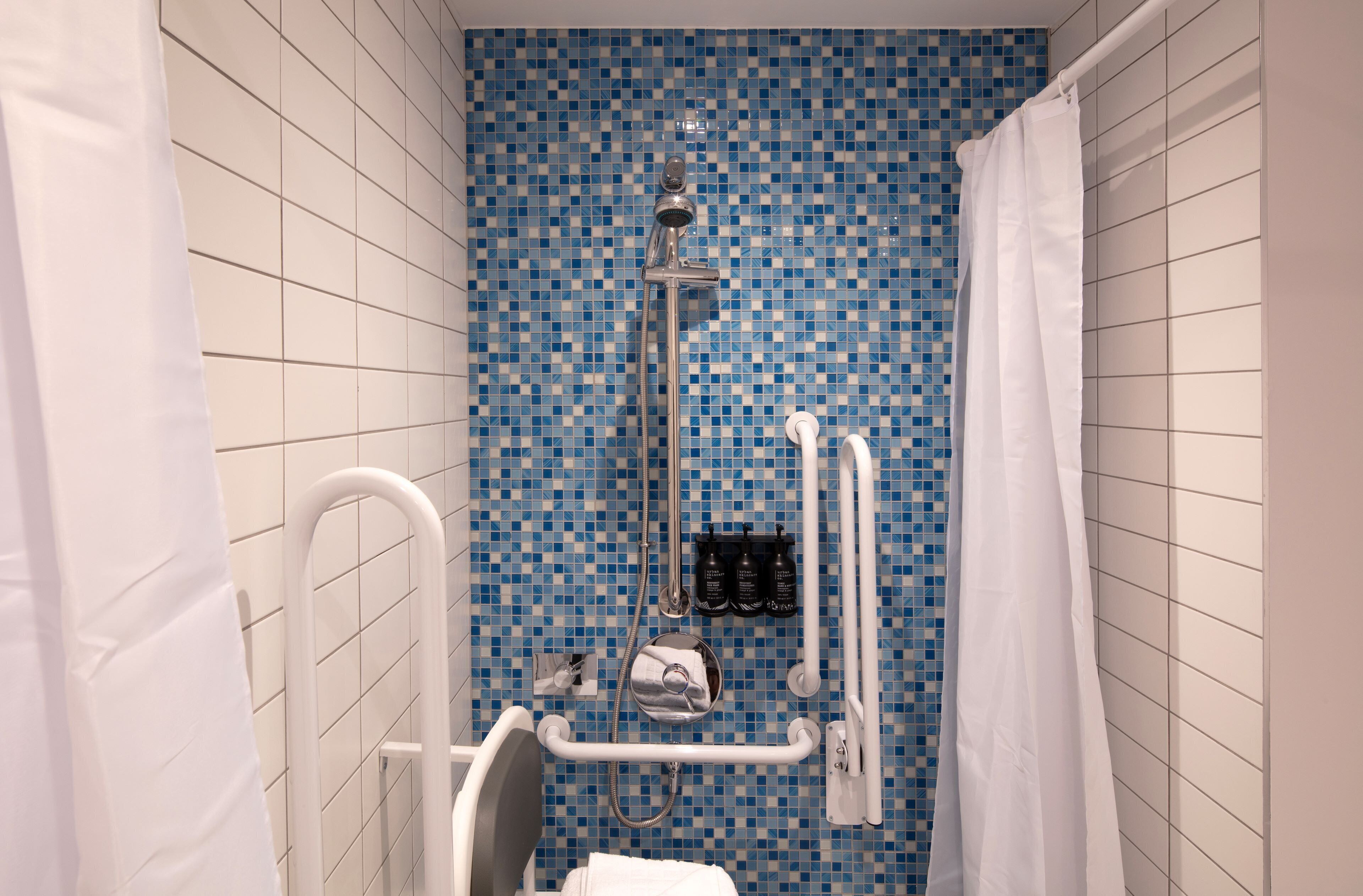 Un baño con pared de mosaicos azules, piso cubierto de baldosas blancas, cortina de ducha blanca y barra de apoyo blanca.