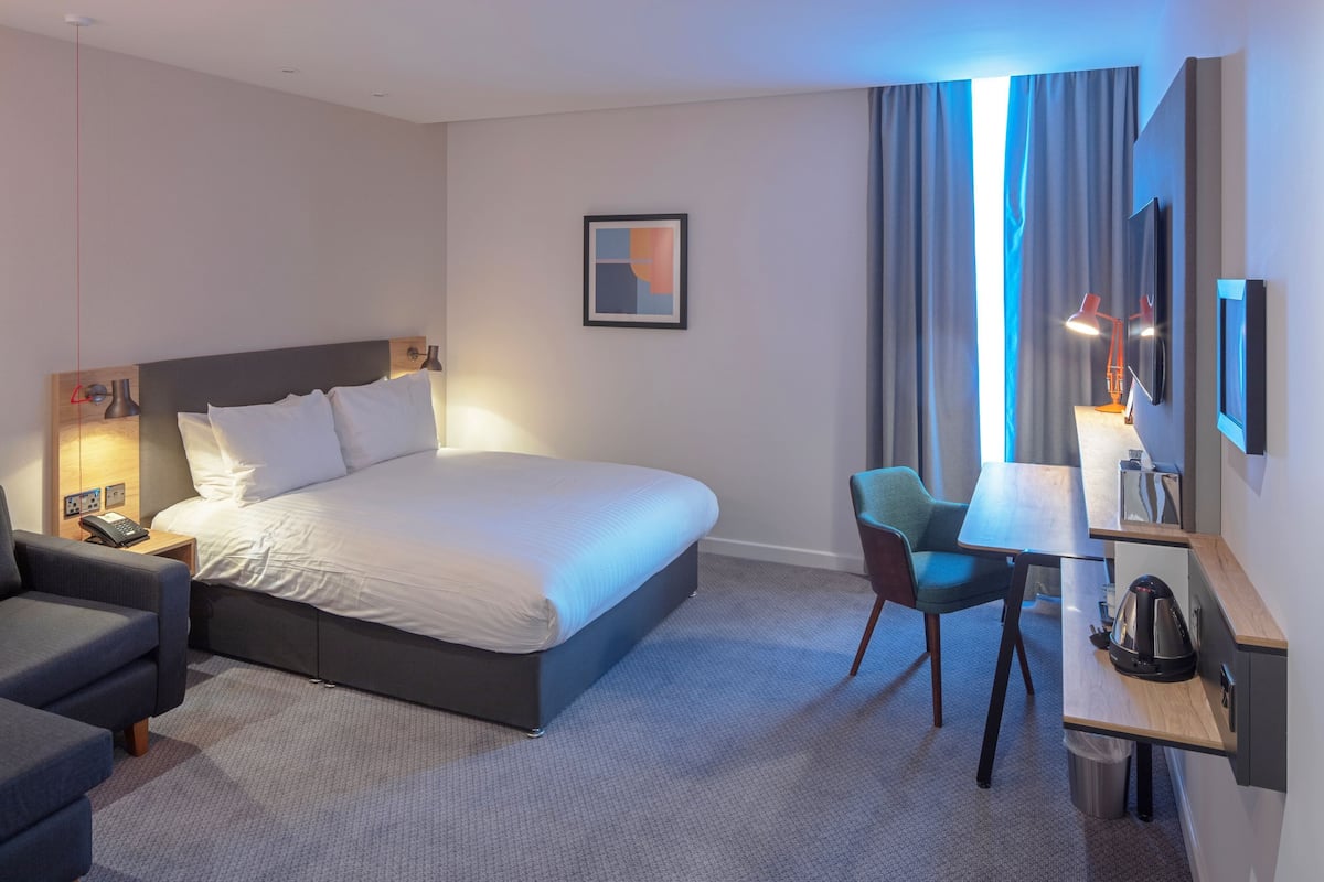 A hotel room with a large bed, a desk with a chair, a TV, and a small kitchenette.