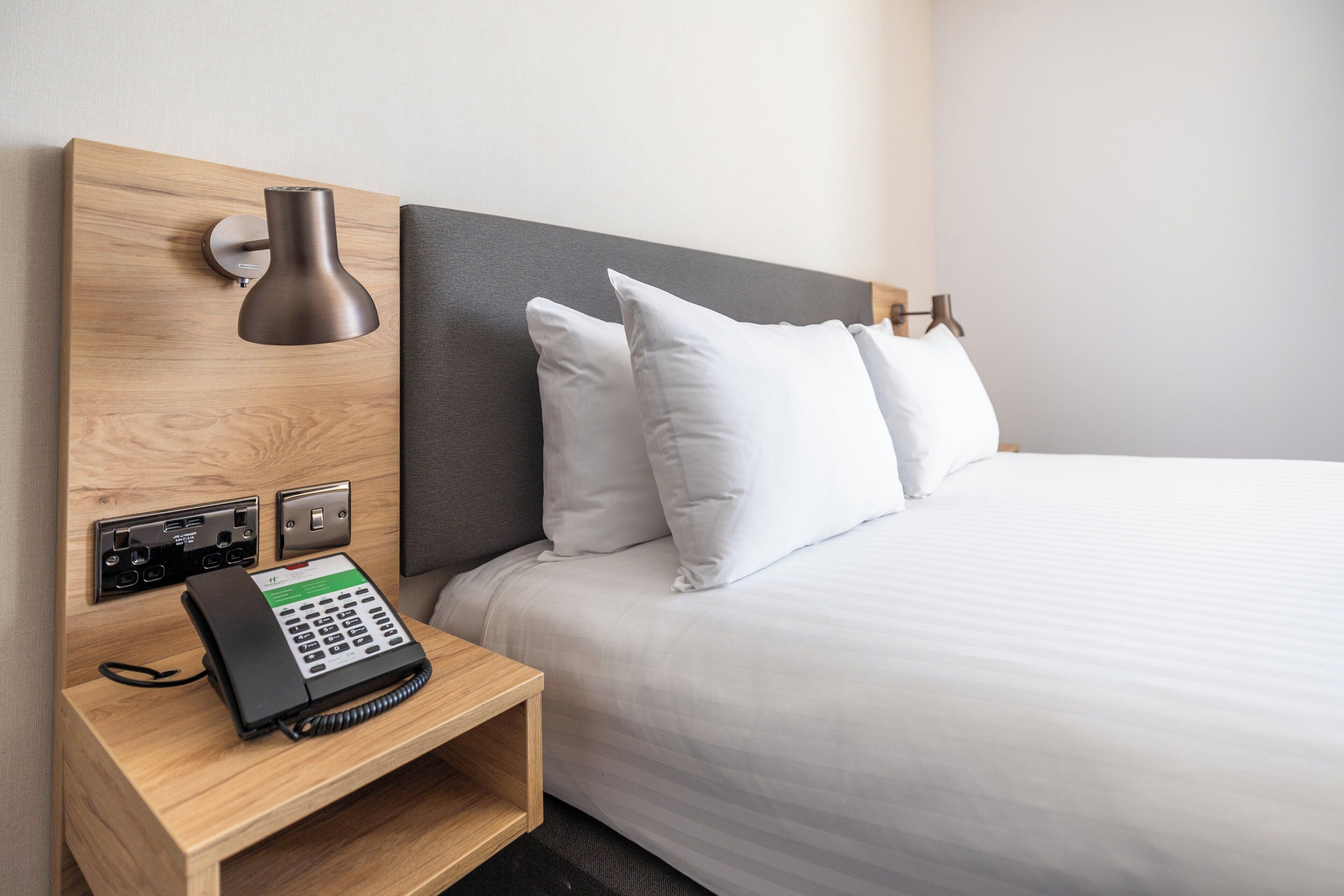 A hotel room with a wooden nightstand, a telephone, a wall-mounted lamp, and a bed with white bedding and pillows.