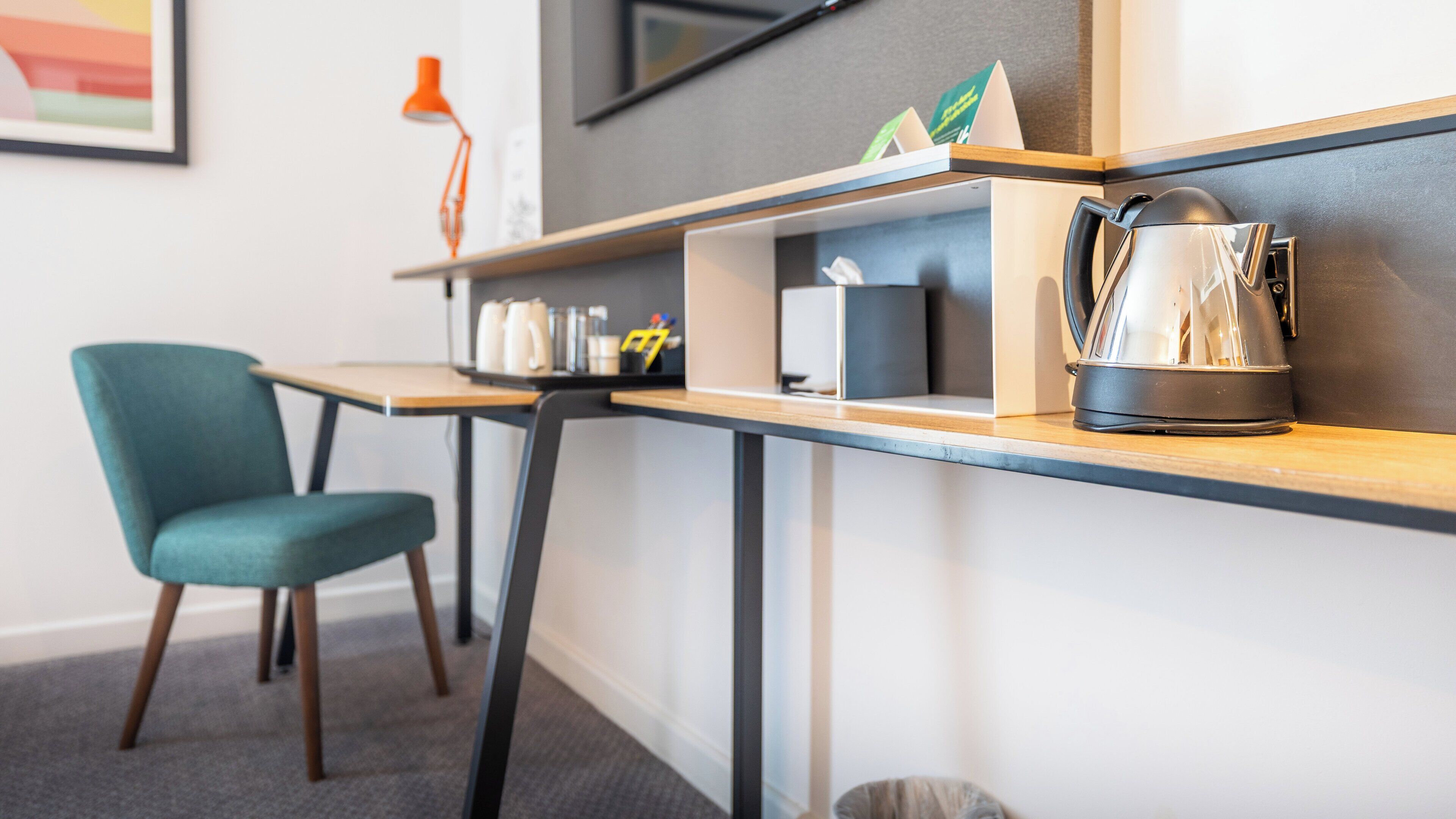 A modern hotel room with a wooden desk, a teal chair, a kettle, and a small shelf with various items.