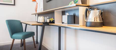 A modern hotel room with a wooden desk, a teal chair, a kettle, and a small shelf with various items.