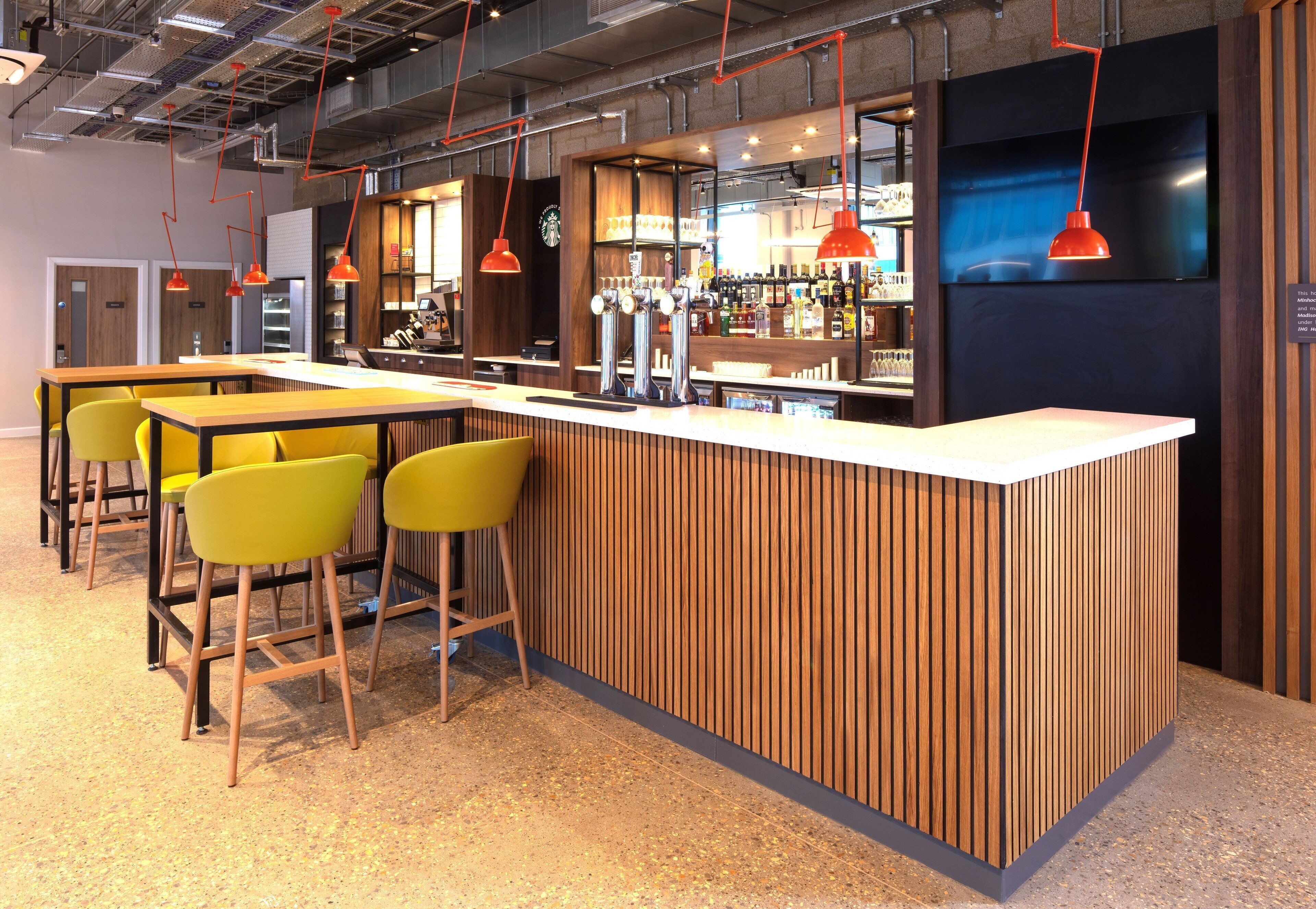 Uma área de bar com balcão de madeira, cadeiras amarelas e luminárias pendentes vermelhas.