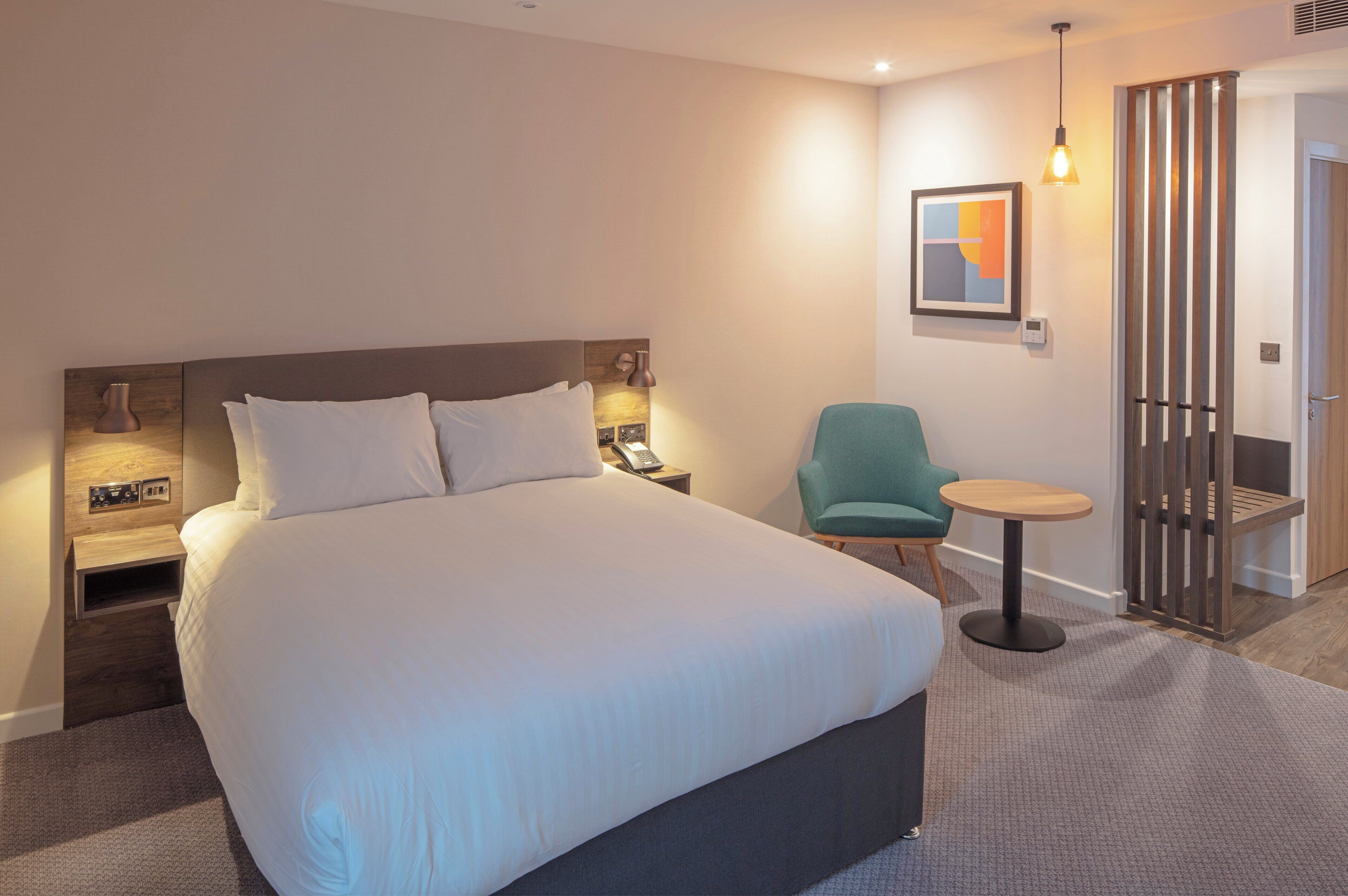 Una habitación de hotel moderna con una cama grande, mesitas de noche con lámparas, una pequeña mesa redonda, un sillón azul turquesa y un panel de madera.