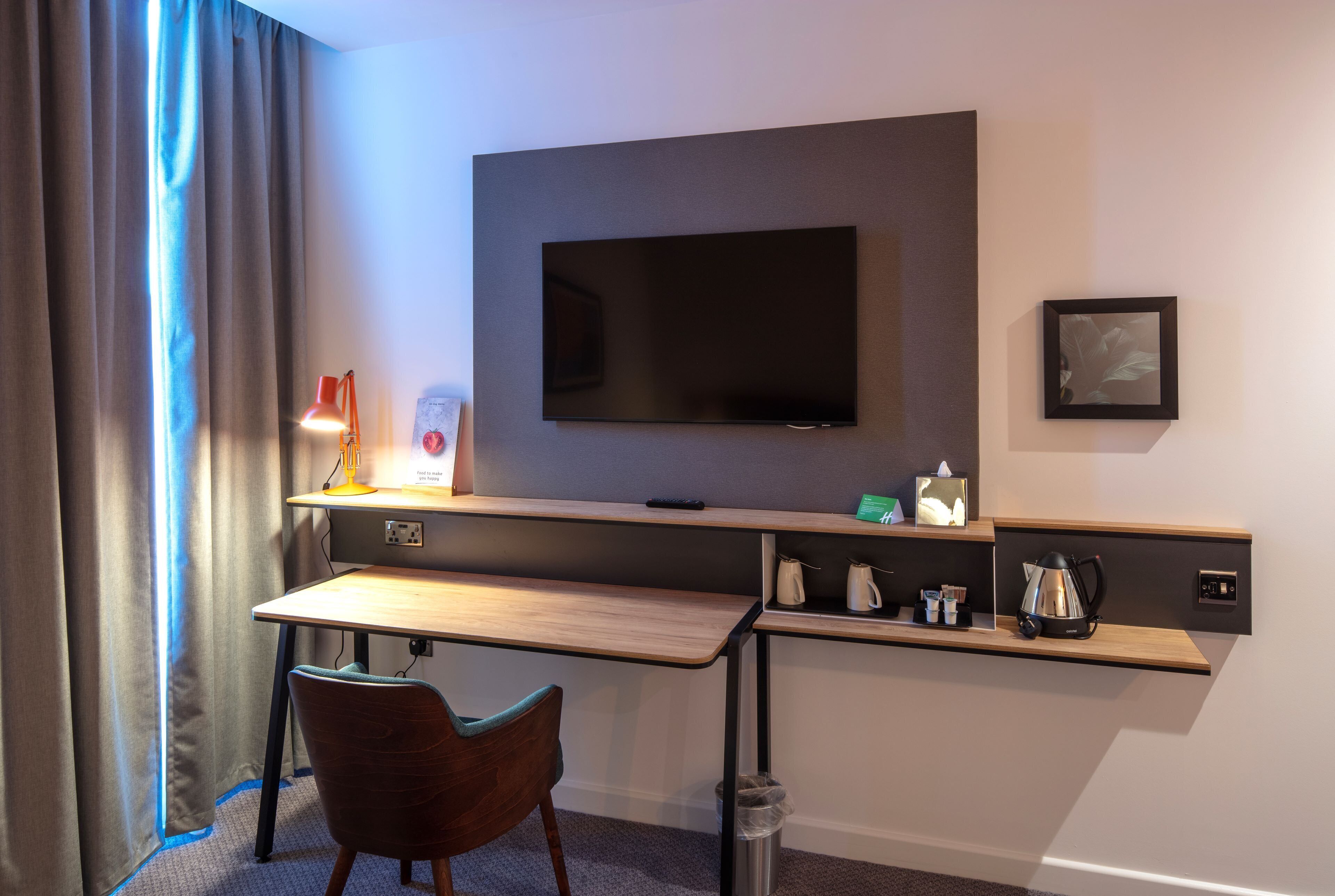 Habitación de hotel con escritorio, silla, televisor y estante con hervidor y otros objetos.