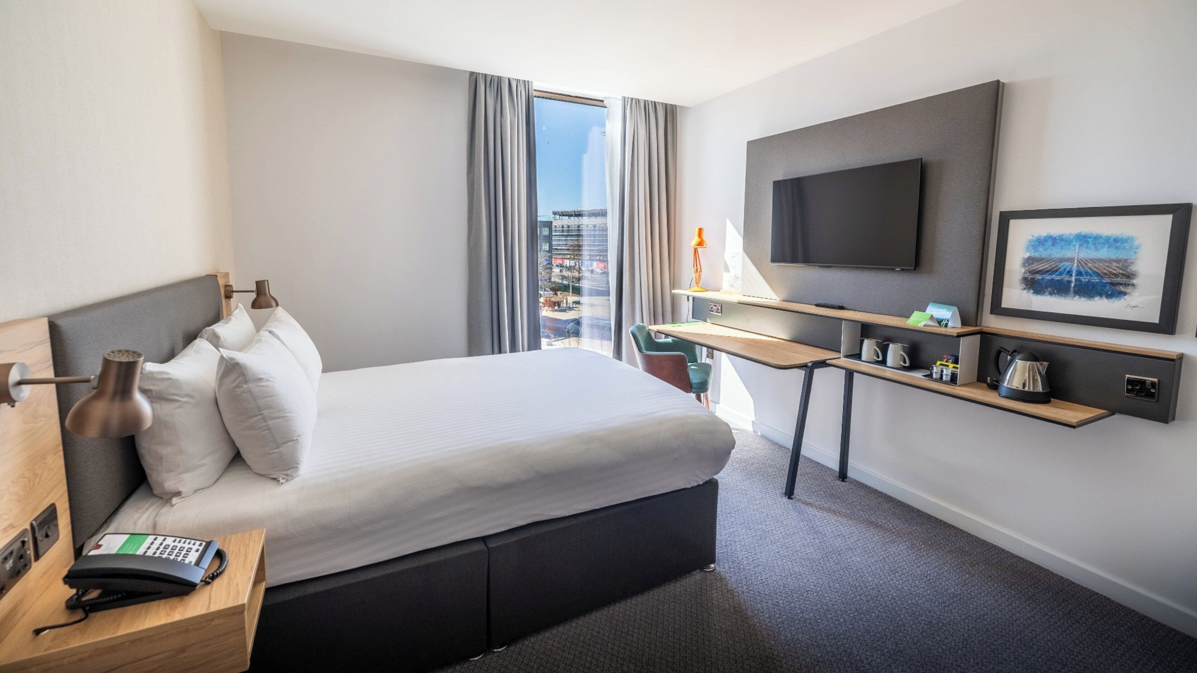 A hotel room with a large bed, a desk with a flat-screen TV, a telephone, and a city view through the window.