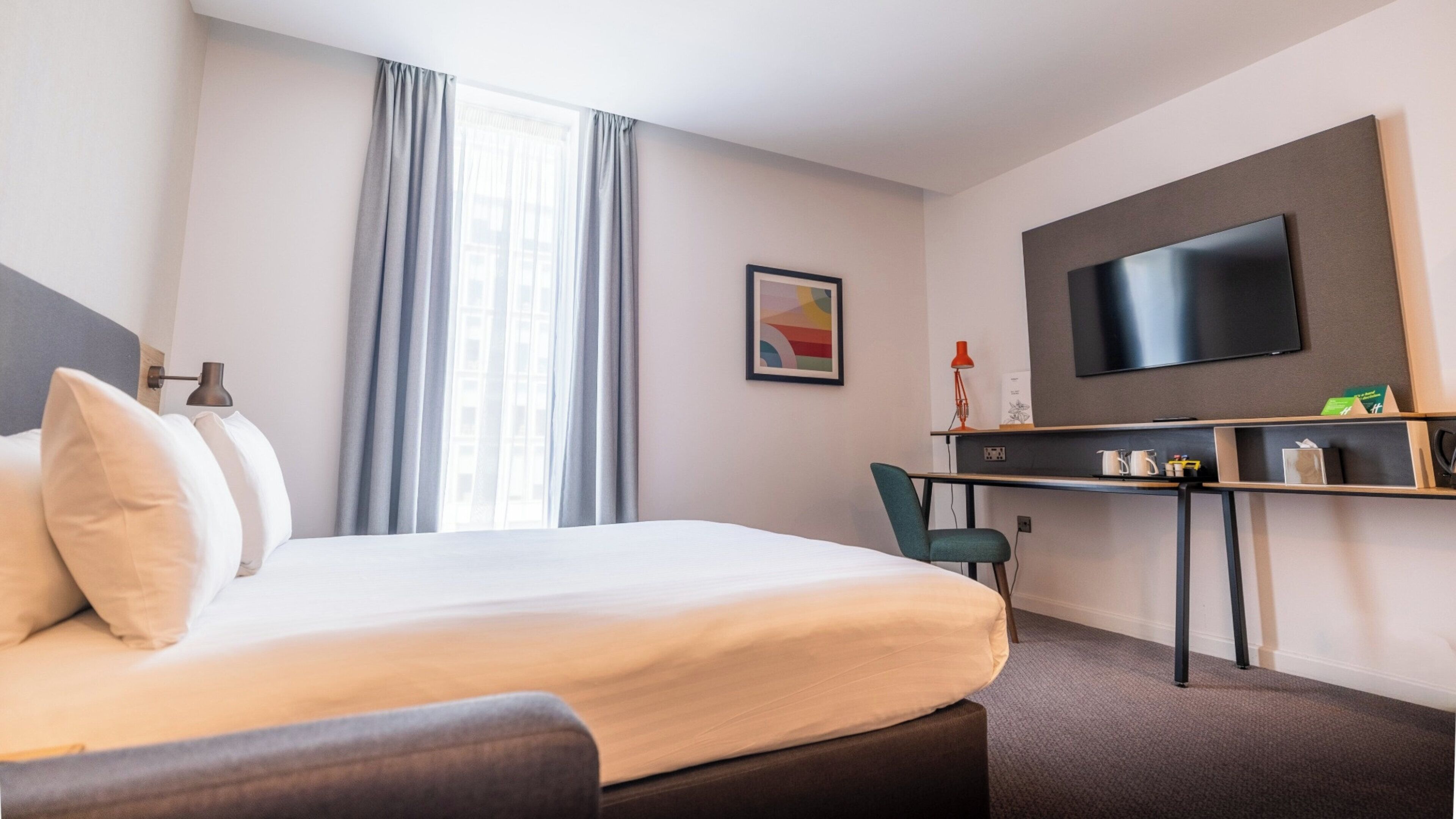 Et hotelværelse med seng, skrivebord, stol, fjernsyn og et vindue med gardiner.
