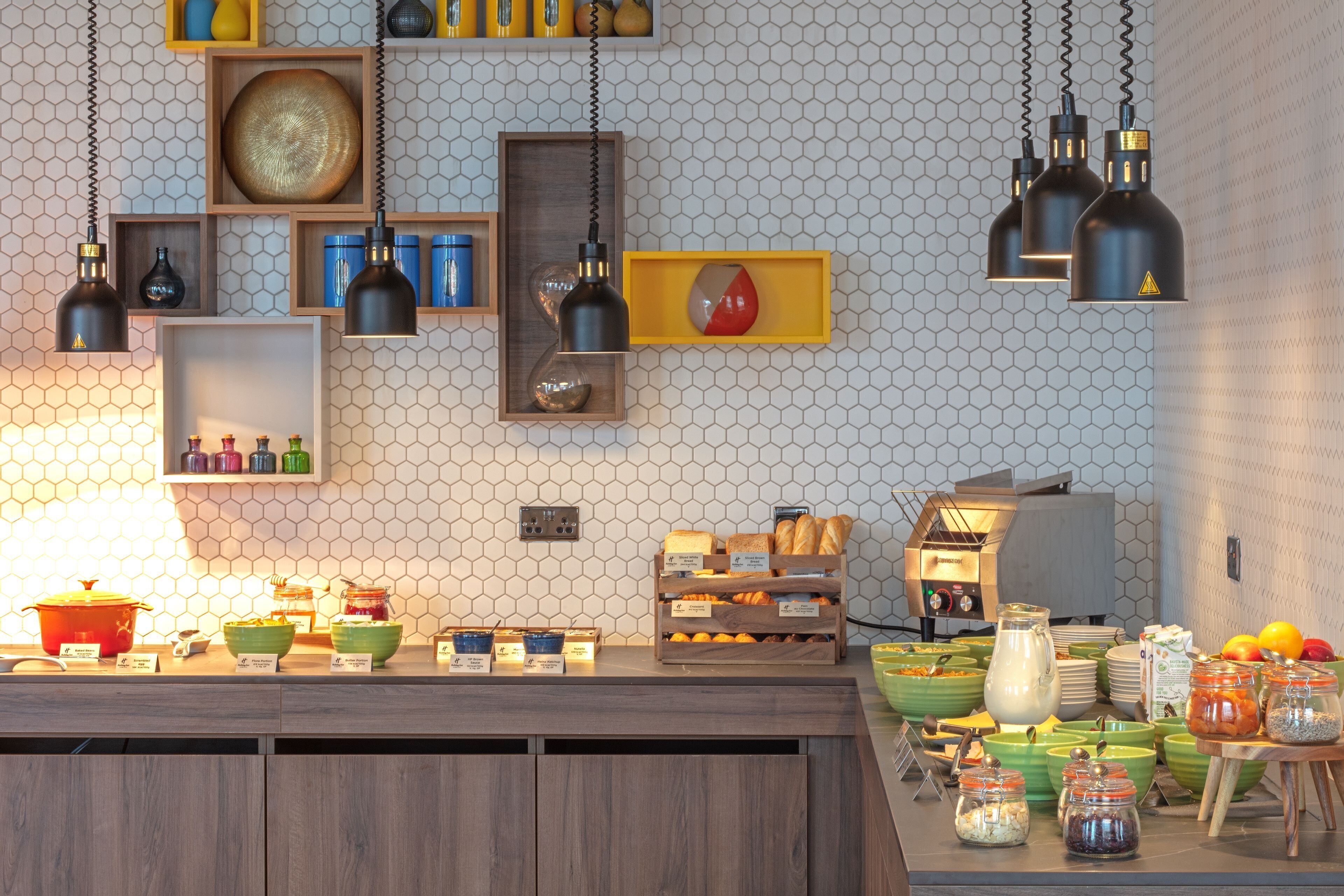 A breakfast buffet with various food items and a modern kitchen area with hanging pendant lights and hexagonal wall tiles.