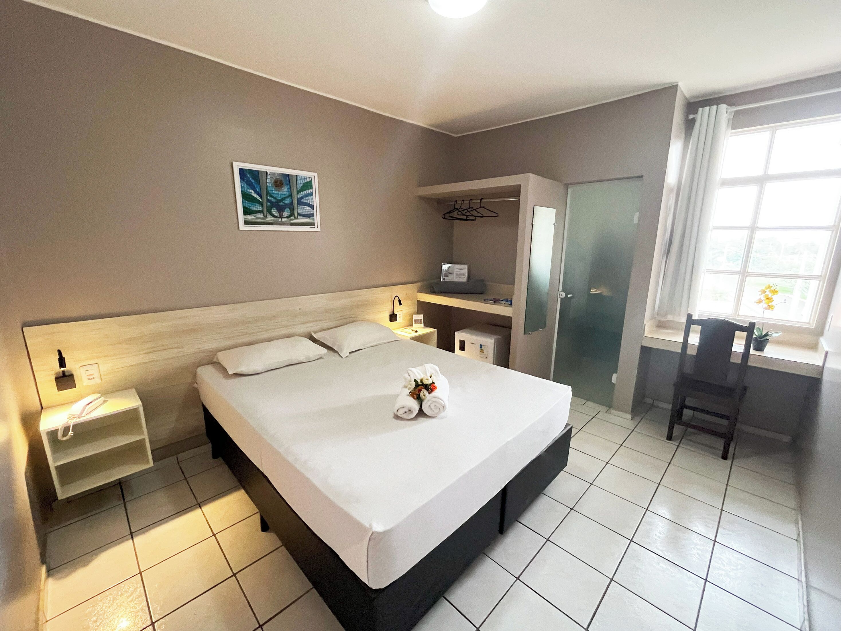 A hotel room with a bed, bedside tables, a desk, a chair, a wardrobe, and a window with curtains.