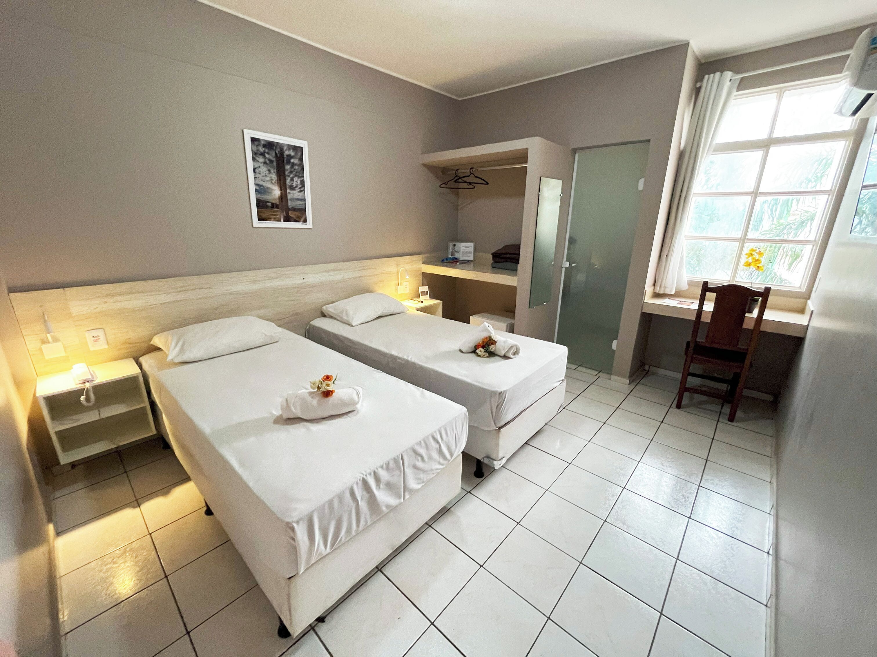 A hotel room with two beds, a desk, a chair, a wardrobe, and a window with a view of greenery.