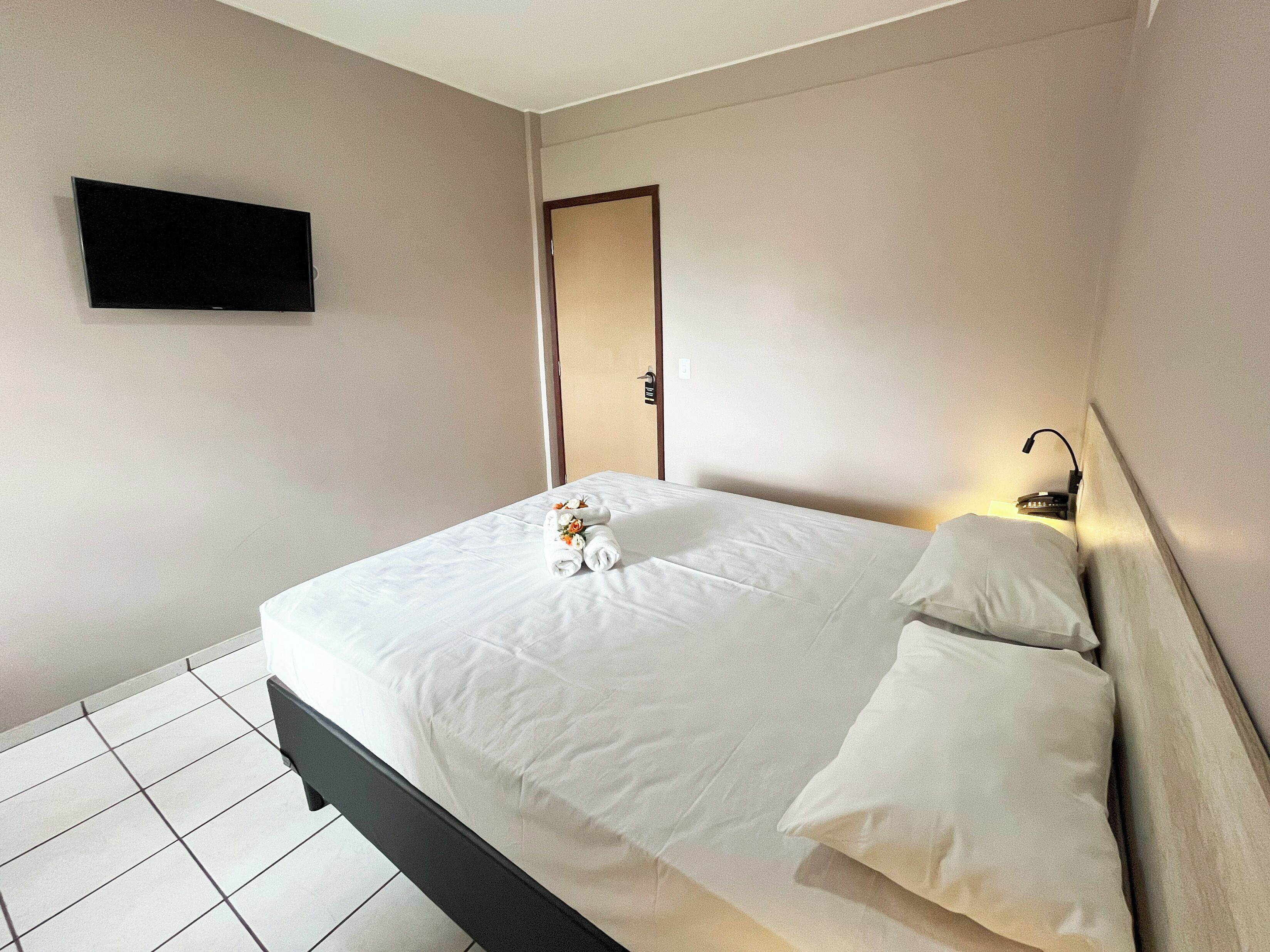 A neatly made bed with white linens and pillows, a flat-screen TV mounted on the wall, and a bedside table with a lamp.