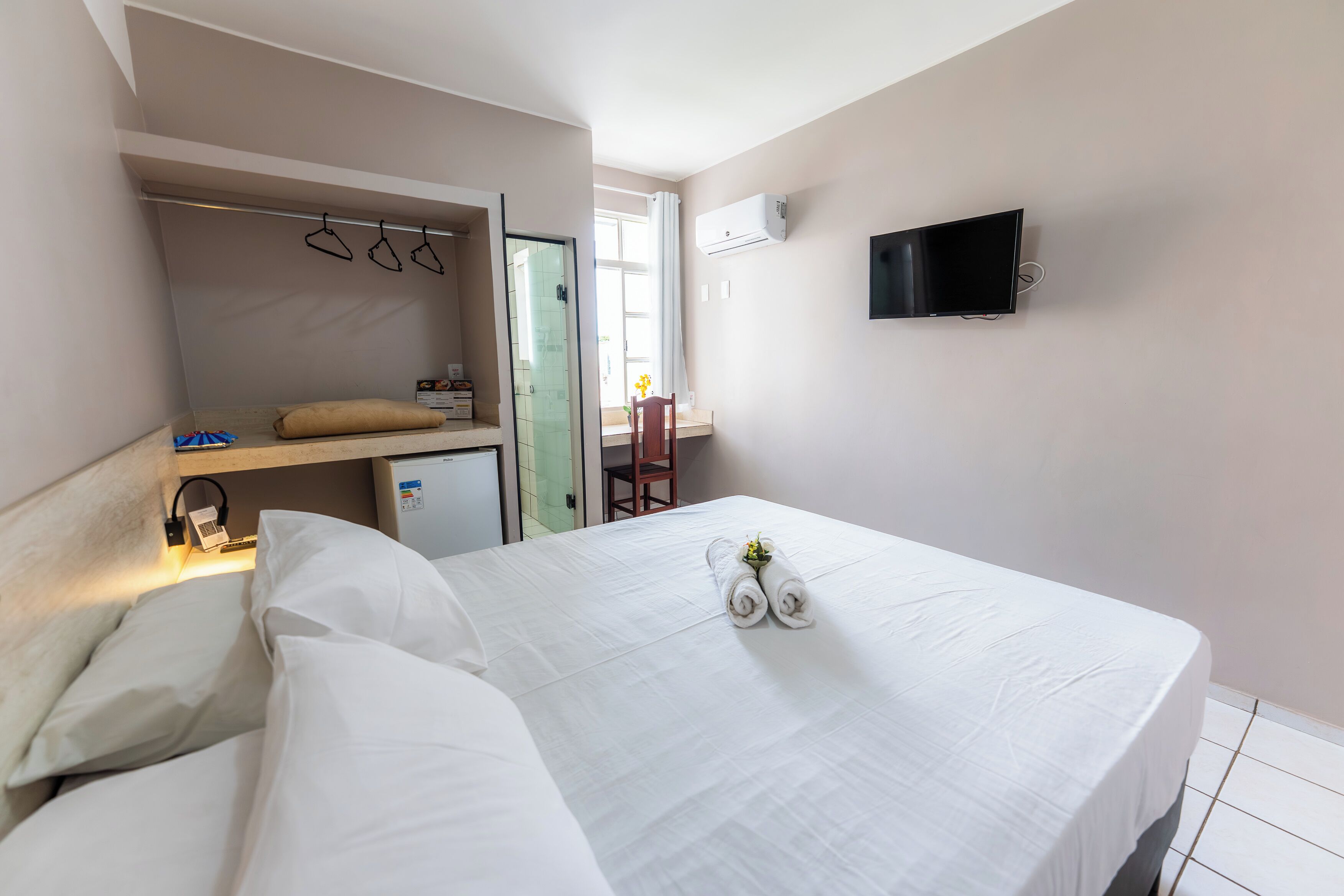Una habitación de hotel con una cama, un televisor montado en la pared, un escritorio pequeño con una silla y una ventana con cortinas.
