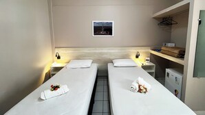 Hotellihuone, jossa on kaksi erillistä sänkyä. Jokaisessa sängyssä on valkoinen peitto ja tyynyjä, pieni yöpöytä, jossa on lamppu, seinälle ripustettu taulu sekä hylly, jossa on pyyhkeitä ja pieni lämmitin.