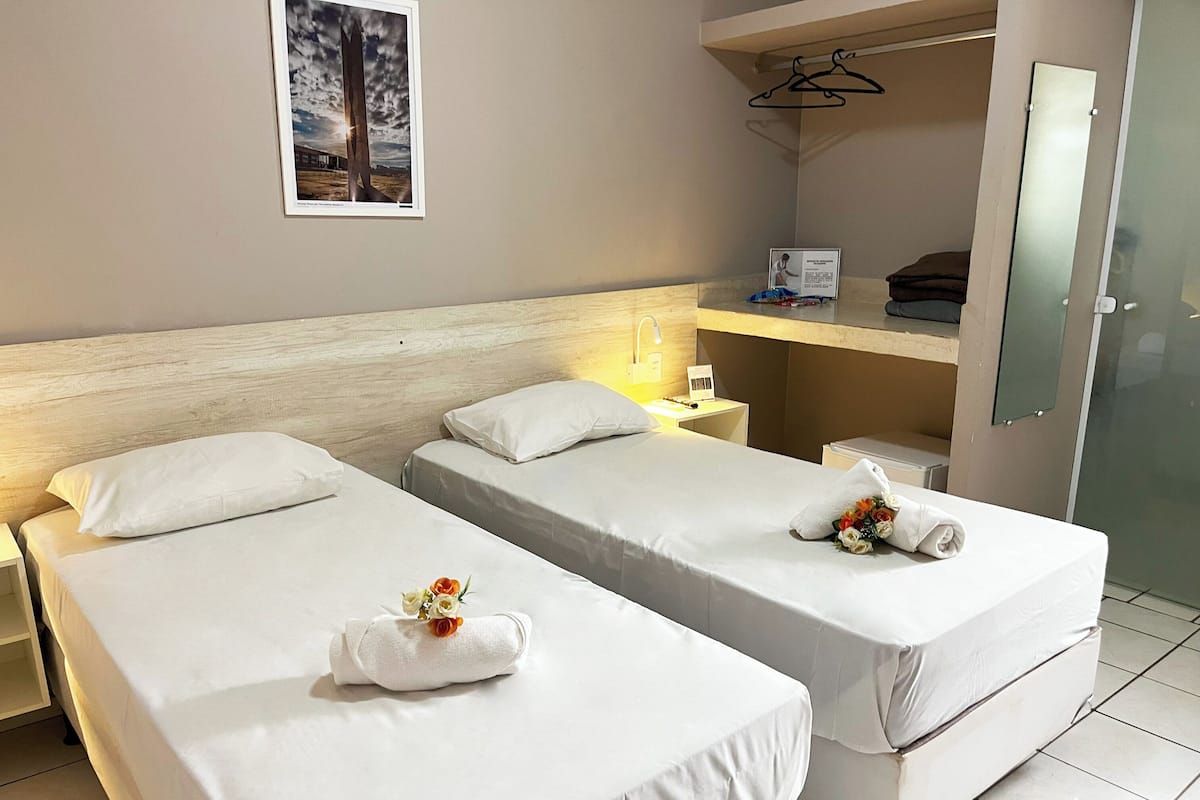 Une chambre d’hôtel avec deux lits simples, une table de chevet avec une lampe, une petite table avec une chaise, un miroir et un tableau encadré au mur.