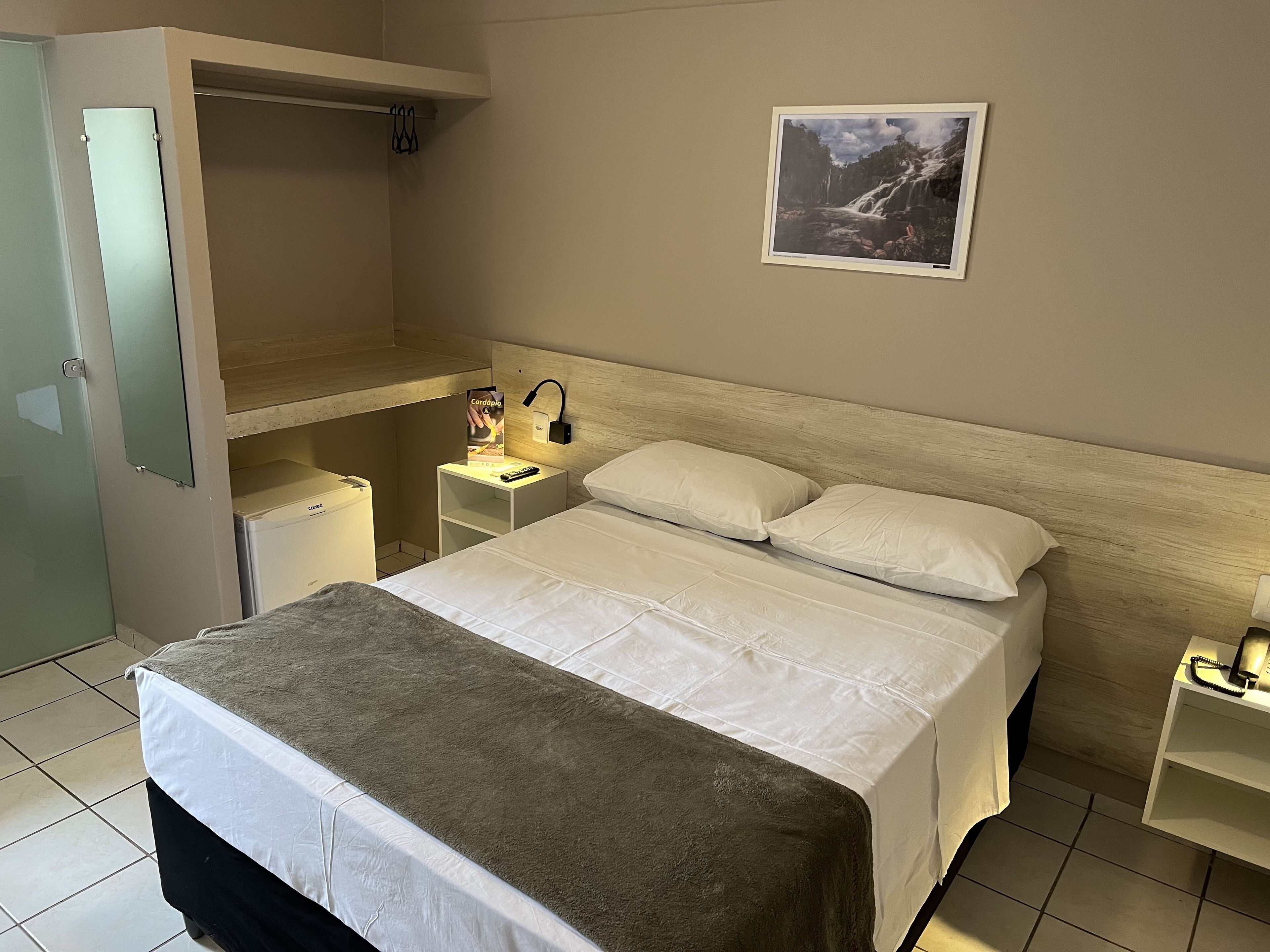 A hotel room with a bed, a nightstand, a bedside table, a wall-mounted picture, and a wardrobe.