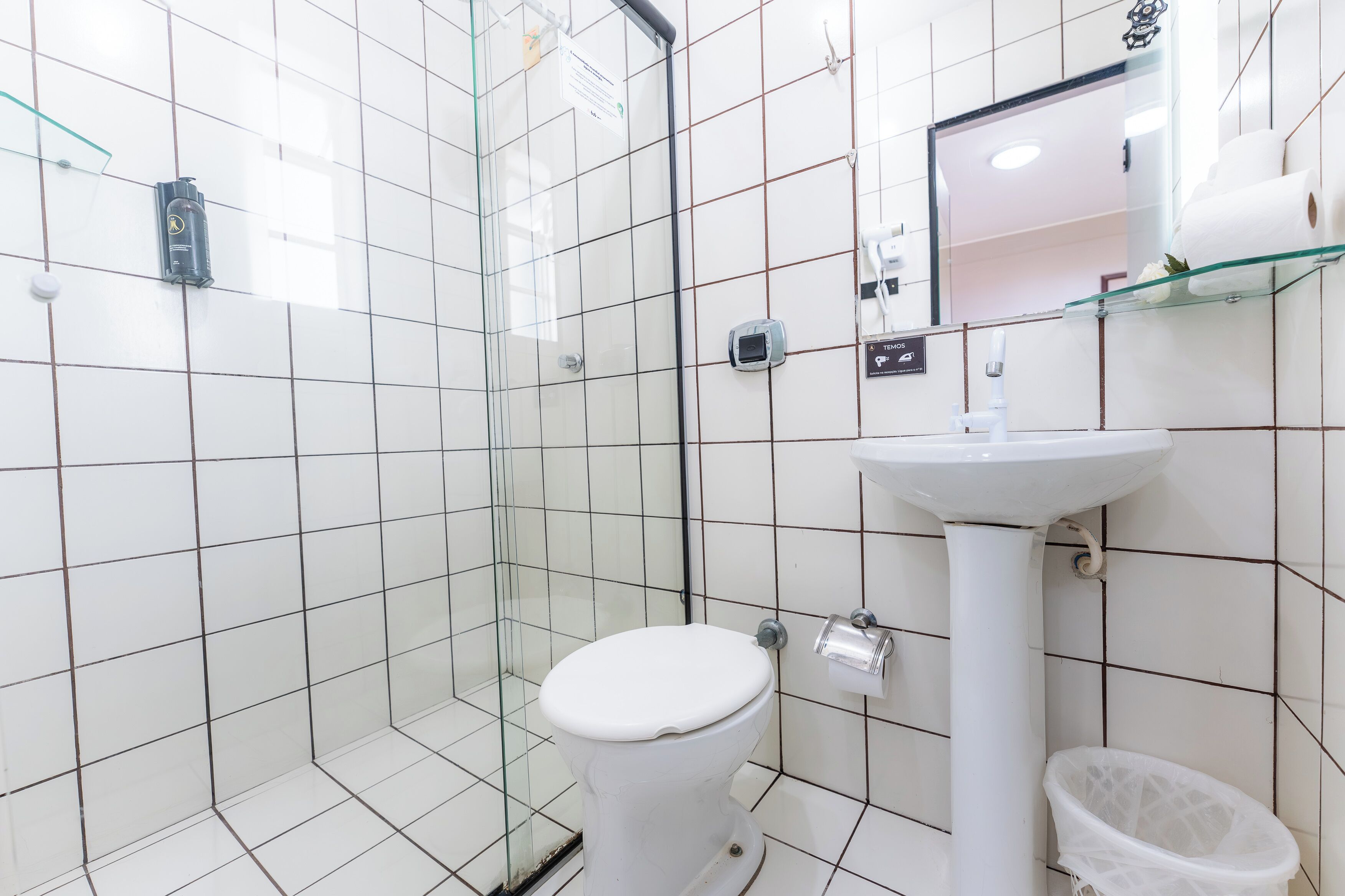 Une salle de bain avec des murs carrelés de blanc, des toilettes blanches, une douche en verre, un lavabo blanc et une étagère en verre où sont rangés les articles de toilette.