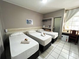 Habitación de hotel con tres camas individuales, un pequeño escritorio con silla, una ventana con cortinas y un calentador fijado a la pared.