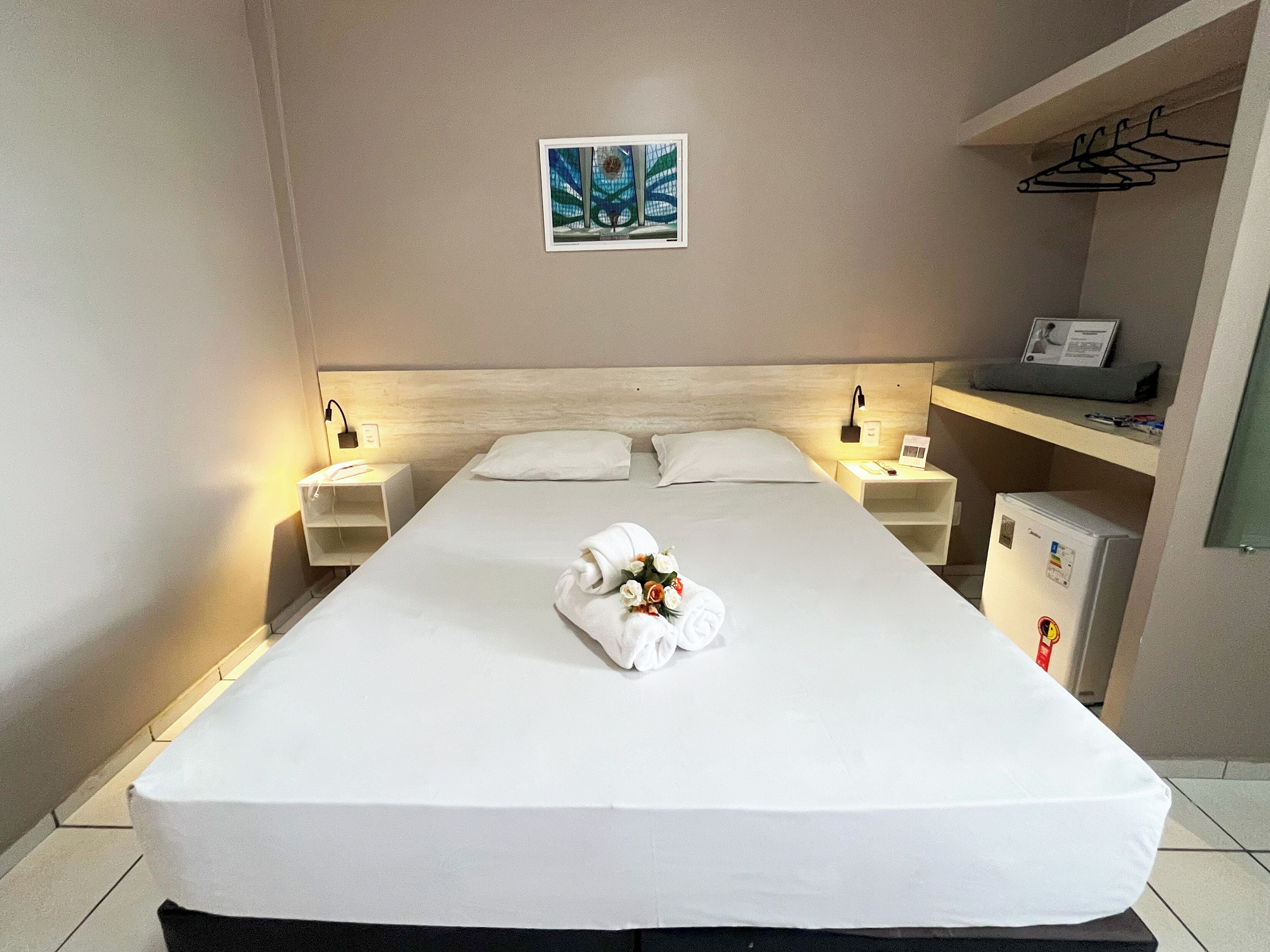 A hotel room with a large bed, two bedside tables, a wall-mounted artwork, a small refrigerator, and a shelf with a hanging garment rack.