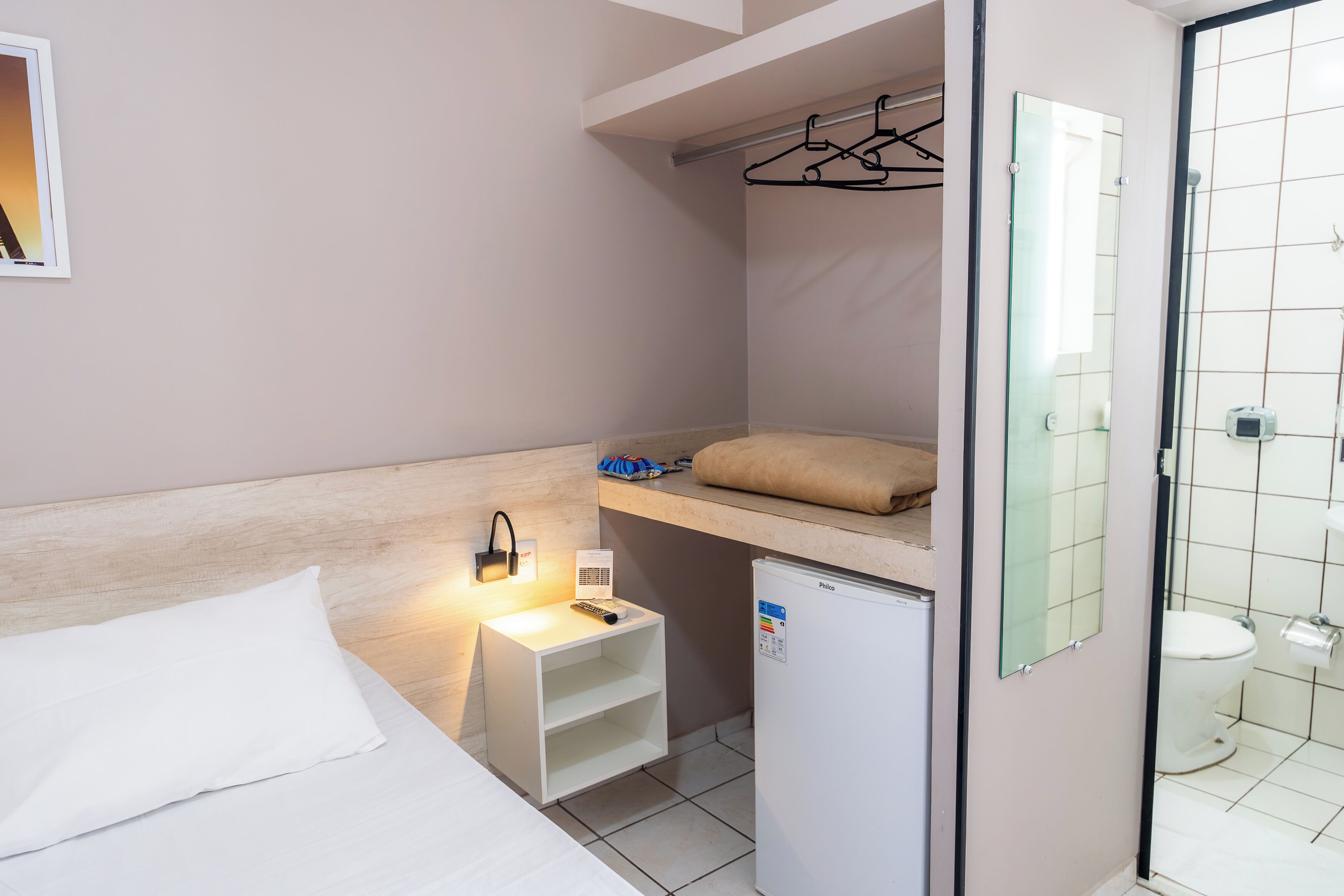 A compact hotel room with a bed, a small bedside table with a lamp, a mini fridge, a shower area, and a hanging clothes rack.