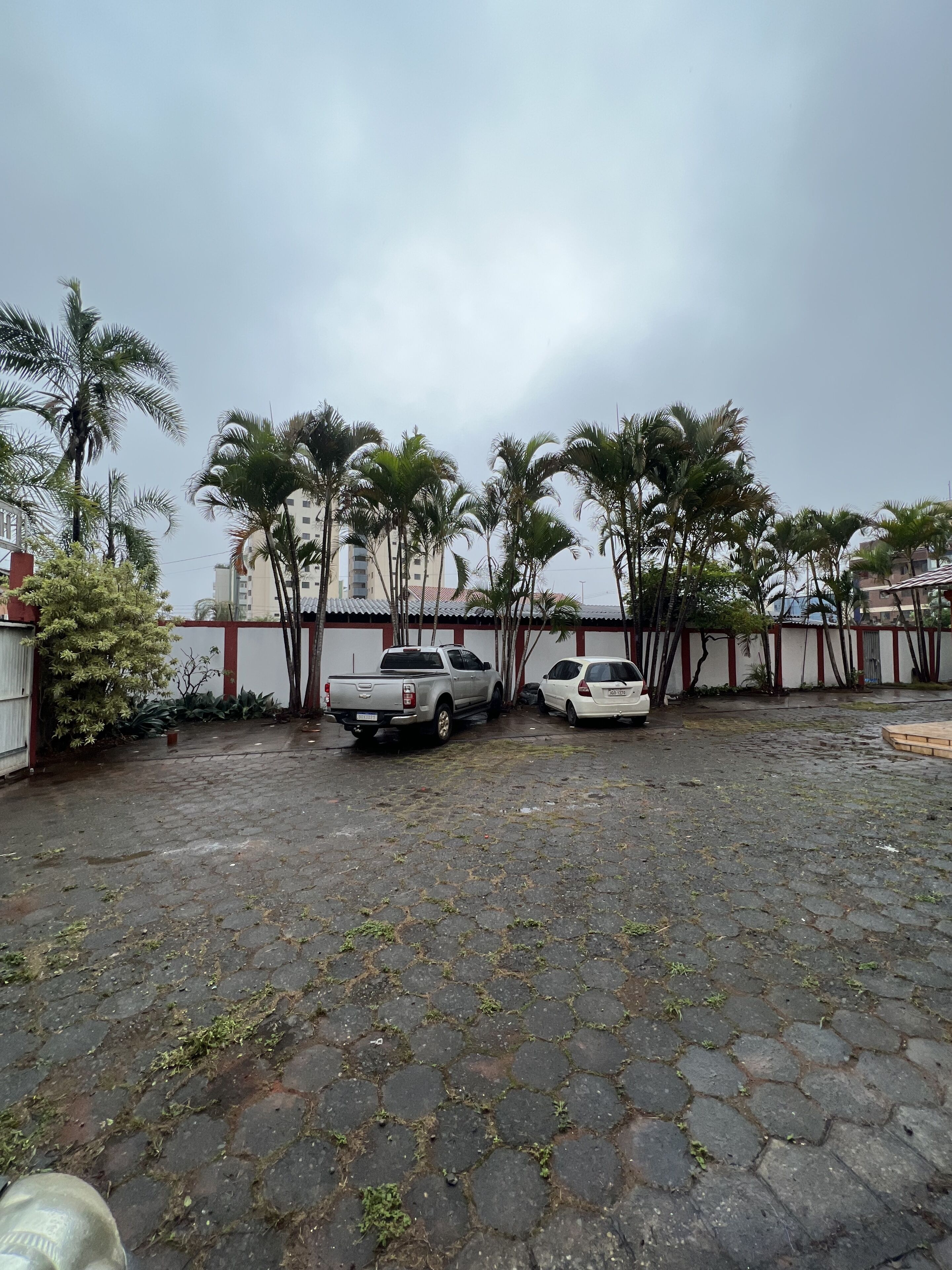 A paved courtyard with a white pickup truck and a white car parked, surrounded by palm trees and buildings in the background.