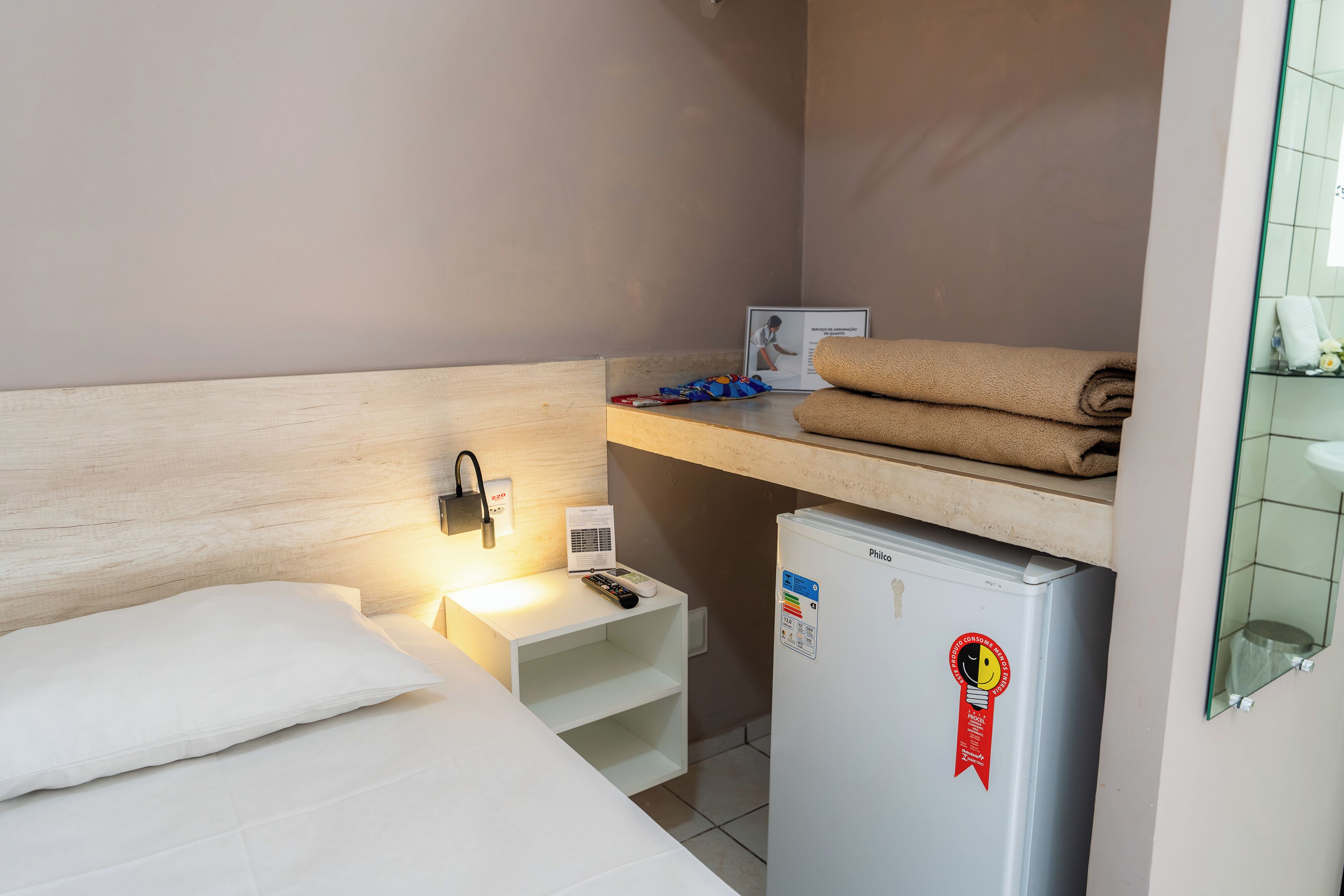 Une chambre d’hôtel compacte comprenant un lit, une petite table de chevet avec une télécommande, un radiateur mural, un réfrigérateur et un porte-serviettes.