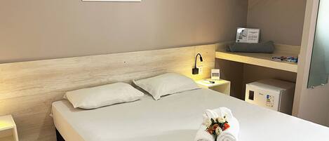 A neatly made bed with pillows, a bedside table with a lamp, a small shelf with items, and a wall art piece.