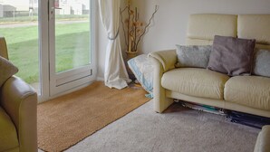 Ein Wohnzimmer mit einem beigefarbenen Sofa, einem kleinen Beistelltisch, einer Vase mit Zweigen und Blick auf eine Rasenfläche durch eine Glasdoor.