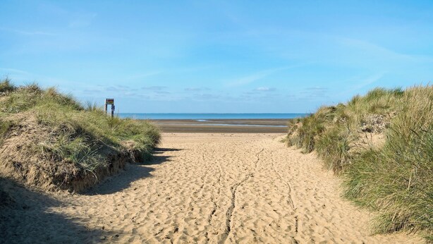 Ein sandiger Küstenpfad führt zum Meer, gesäumt von grasbewachsenen Dünen.
