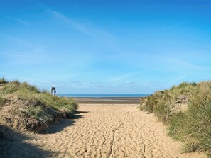 En sti af sand der fører ned til havet, omkranset af græsbeklædte klitter.