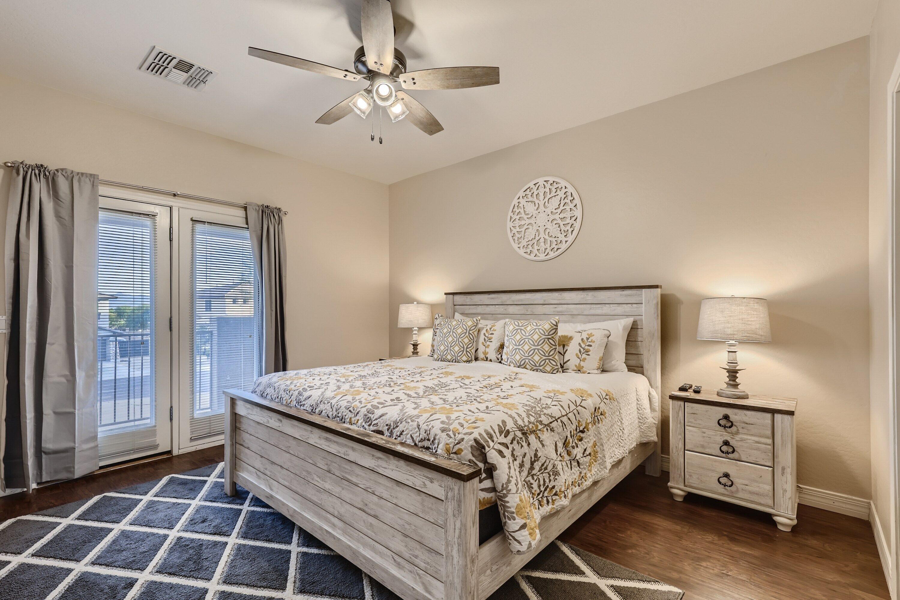 A bedroom with a wooden bed, a nightstand with a lamp, a ceiling fan, and a view through the window.
