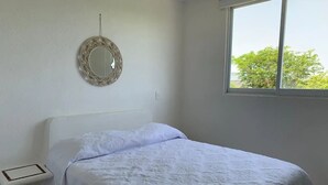 Un dormitorio con cama, mesita de noche, espejo en la pared y ventana con vista a árboles.