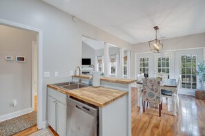 Una cucina moderna con piano di lavoro in legno, lavello in acciaio inossidabile e lavastoviglie. C'è un'area pranzo con tavolo e sedie, e un'ampia finestra che permette l'ingresso di luce naturale.