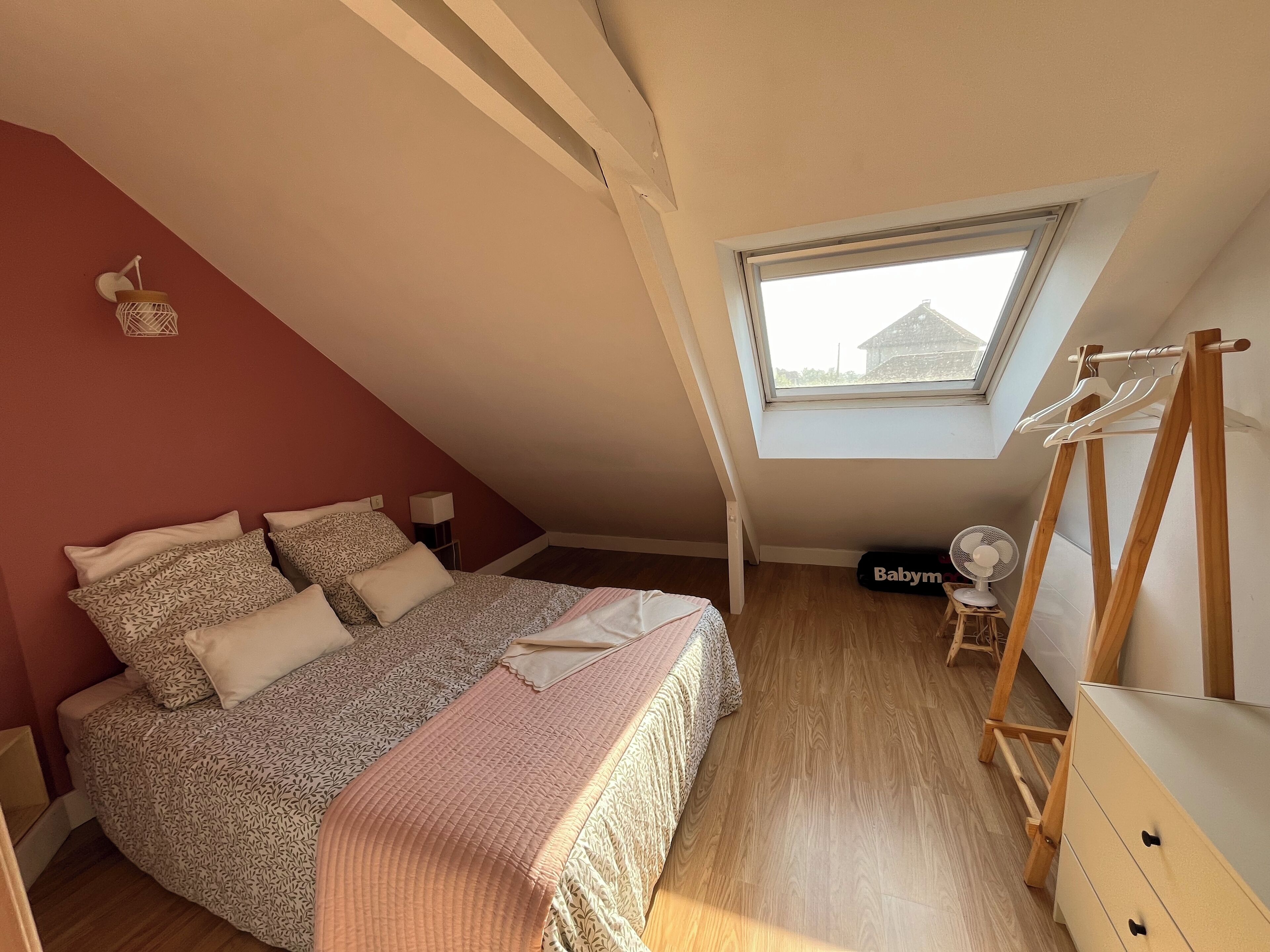 A bedroom with a sloped ceiling, a skylight, a bed with patterned bedding, a bedside table, a dresser, and a hanging clothes rack.
