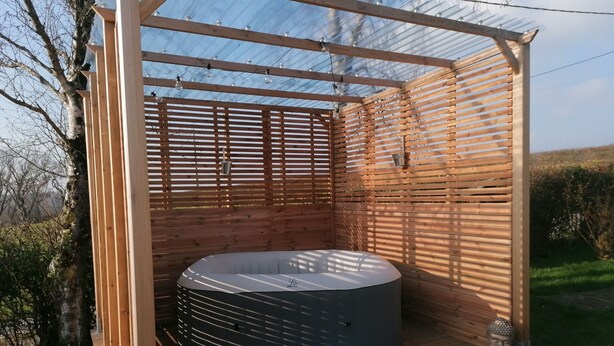 A wooden hot tub structure with a transparent roof, surrounded by wooden slats, and a blue container on the deck.