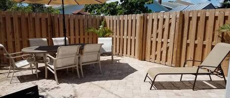 Un patio con cerramiento de madera, una mesa redonda con sillas, una tumbona y un gran paraguas.